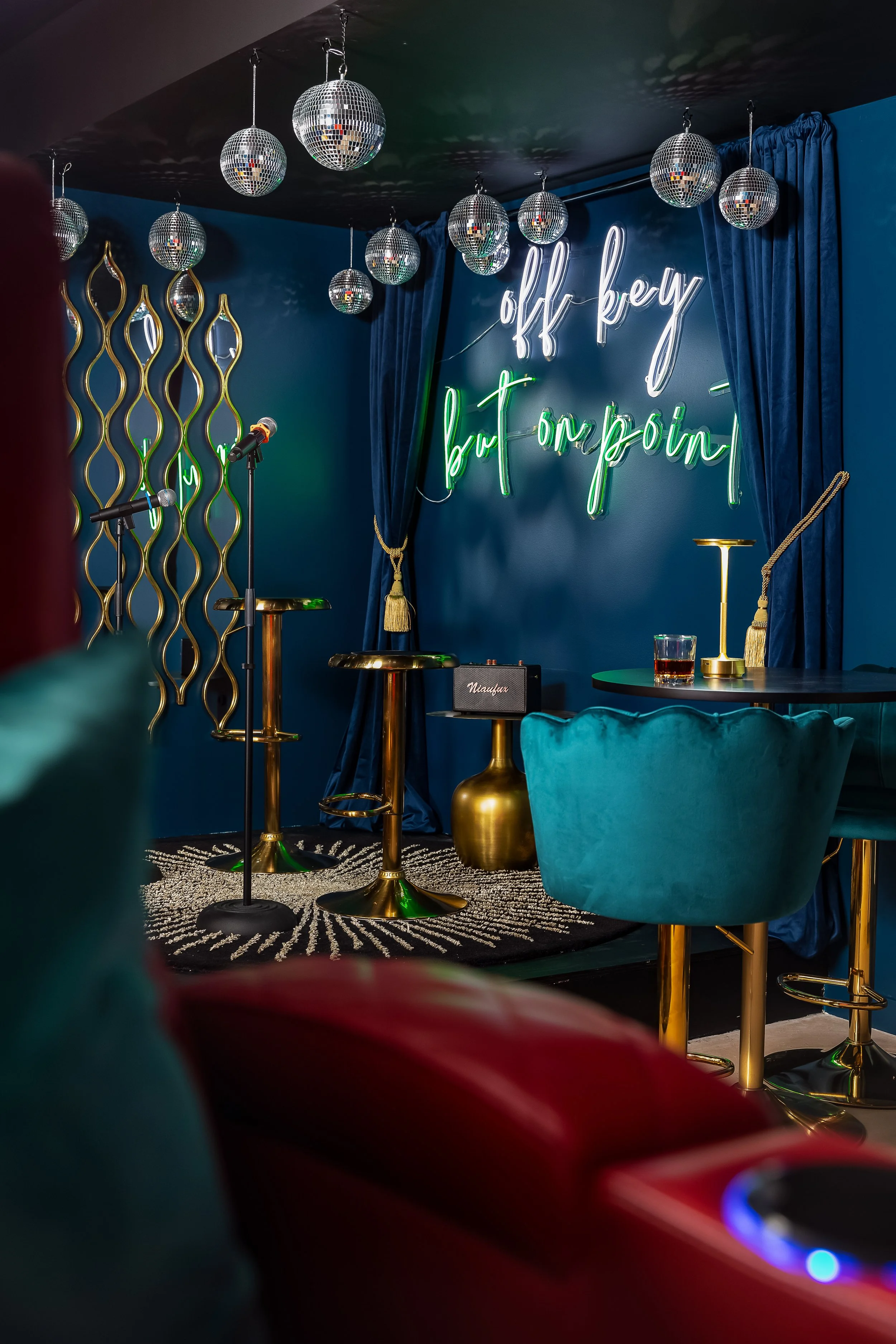Stage with microphone stands, a small amplifier, a candle holder, and a glass of drink, set against dark blue walls with a neon sign that reads 'off key but still popin', surrounded by blue curtains, gold accents, and disco balls hanging from the cei