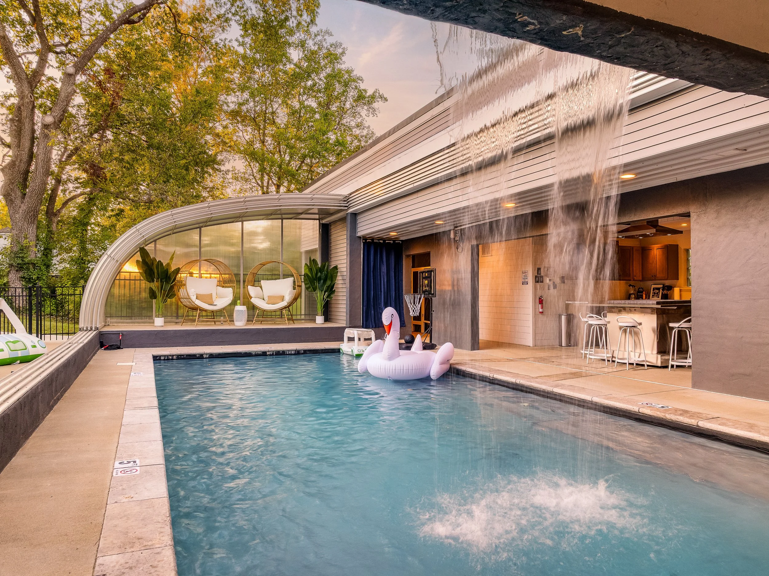 Indoor swimming pool with a large inflatable swan float, water fountain feature, and seating area with hanging egg chairs and potted plants in the background.