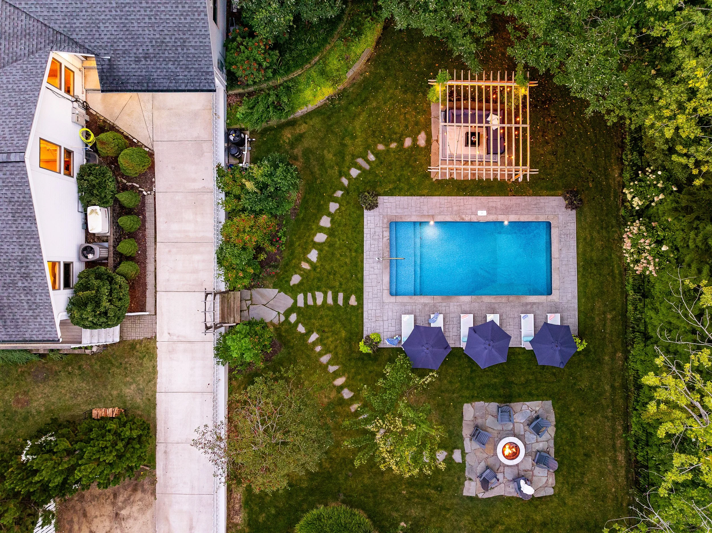 Aerial view of a backyard with a swimming pool, grassy areas, outdoor seating with umbrellas, a fire pit, a small wooden structure, and surrounding trees.