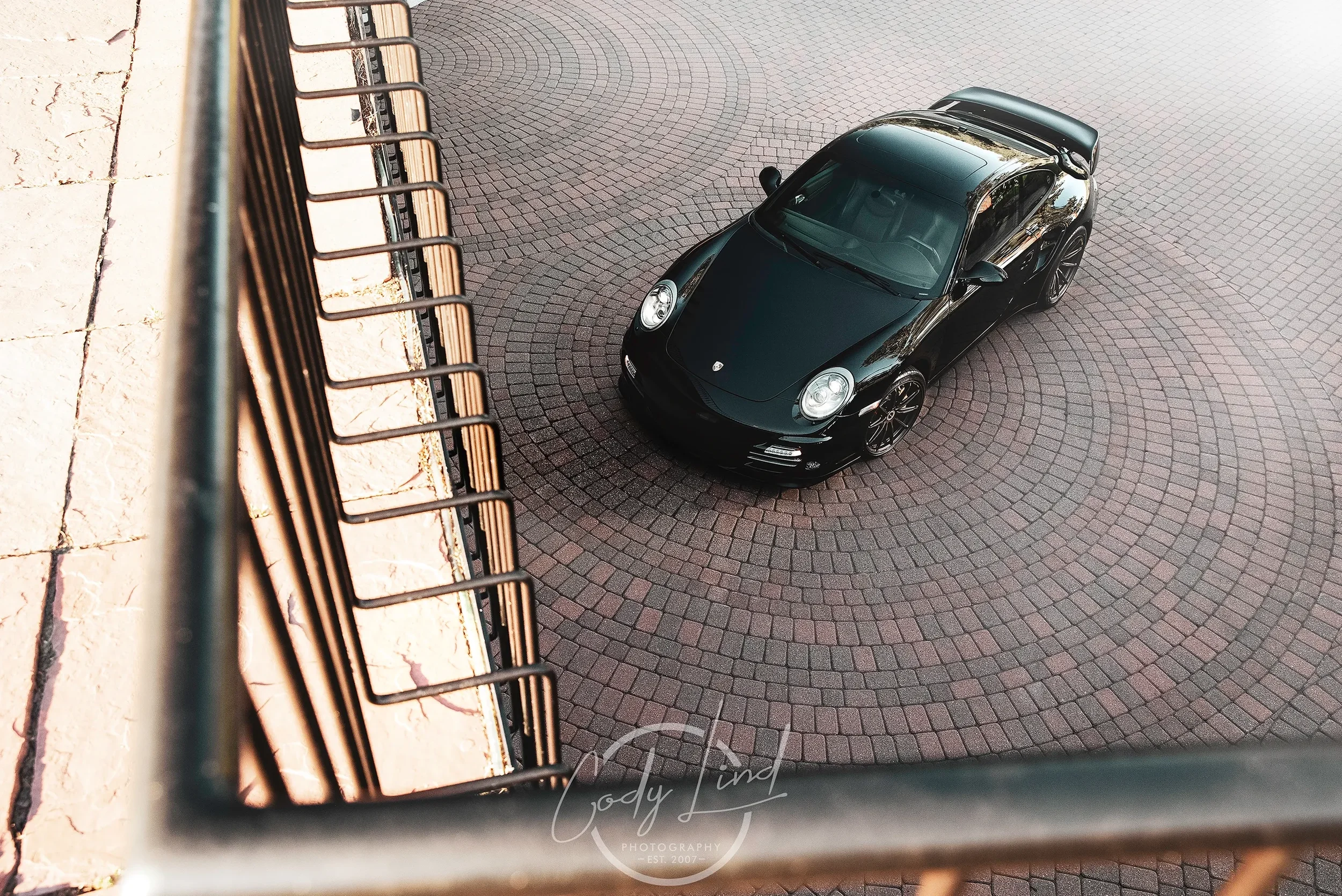 A black Porsche sports car parked on a circular brick driveway seen from above, with part of a metal railing and beige stone wall in the foreground.