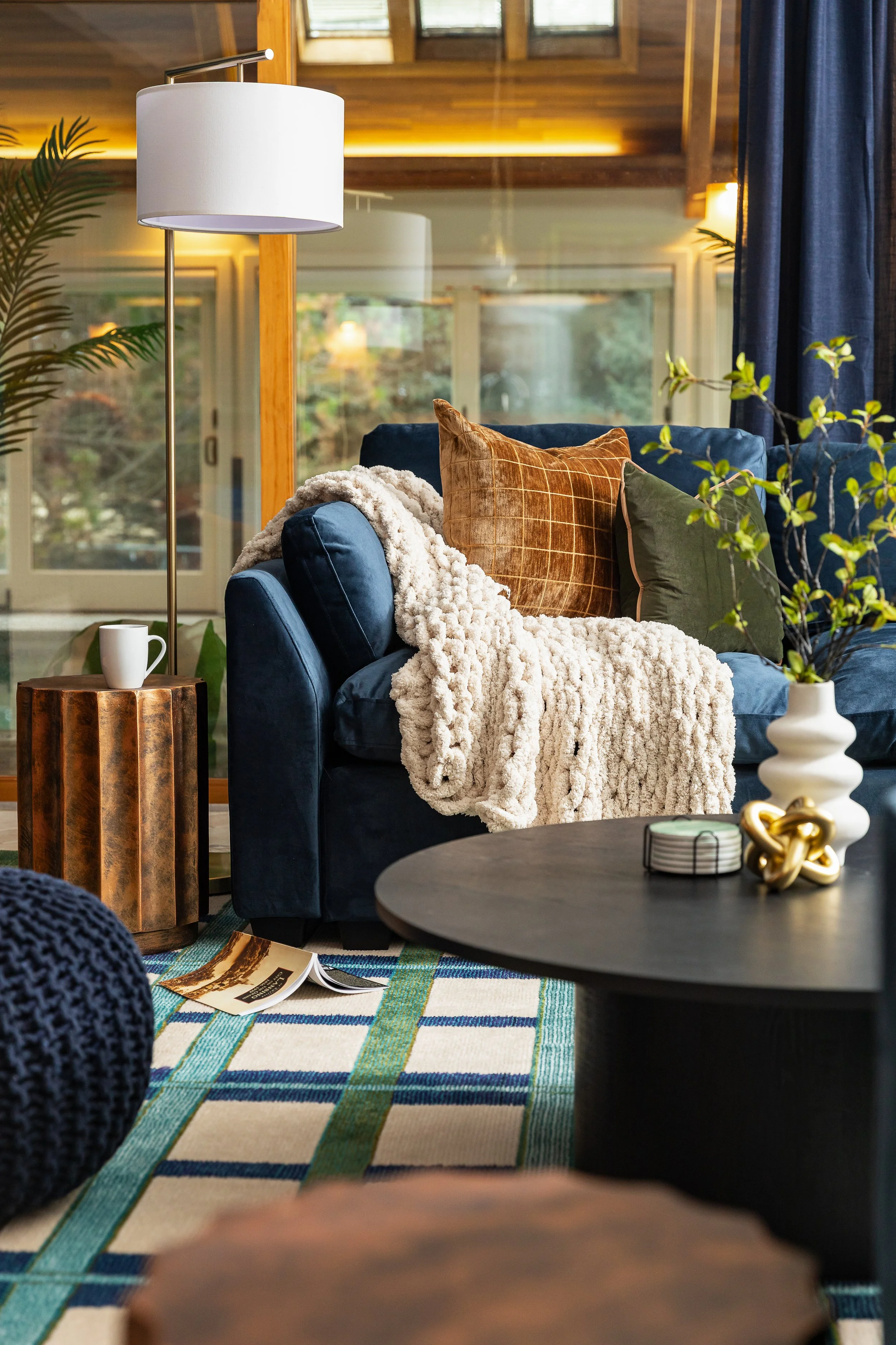 A cozy living room with a blue velvet sofa, throw pillows, a cream fluffy blanket, a wooden side table with a white mug, a modern black coffee table with decorative objects, a white vase with green branches, a colorful striped rug, and large windows 