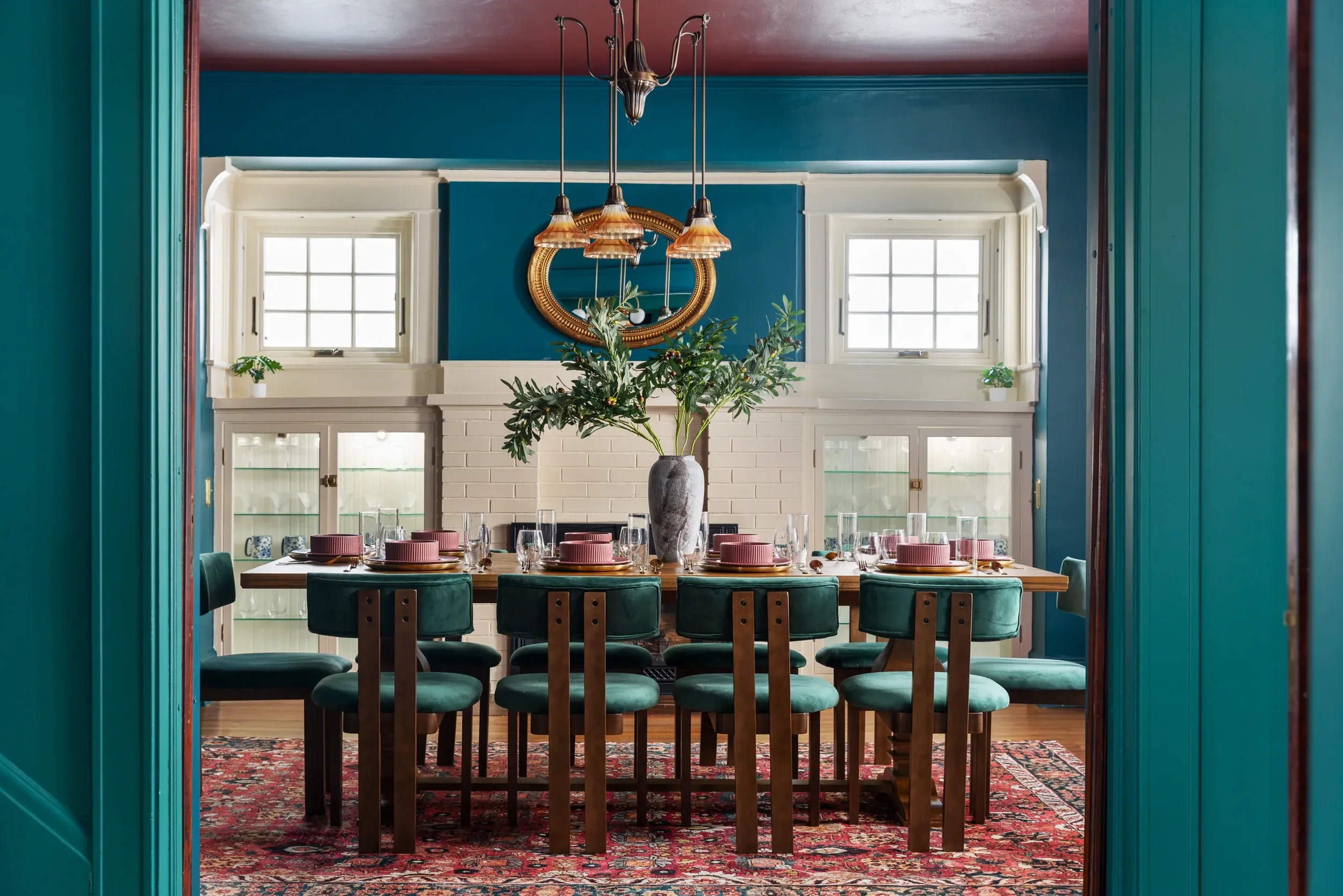 A dining room viewed through an open doorway, featuring a large table set with pink plates, glassware, and a vase with greenery, surrounded by green chairs, with a blue and white interior design and windows in the background.