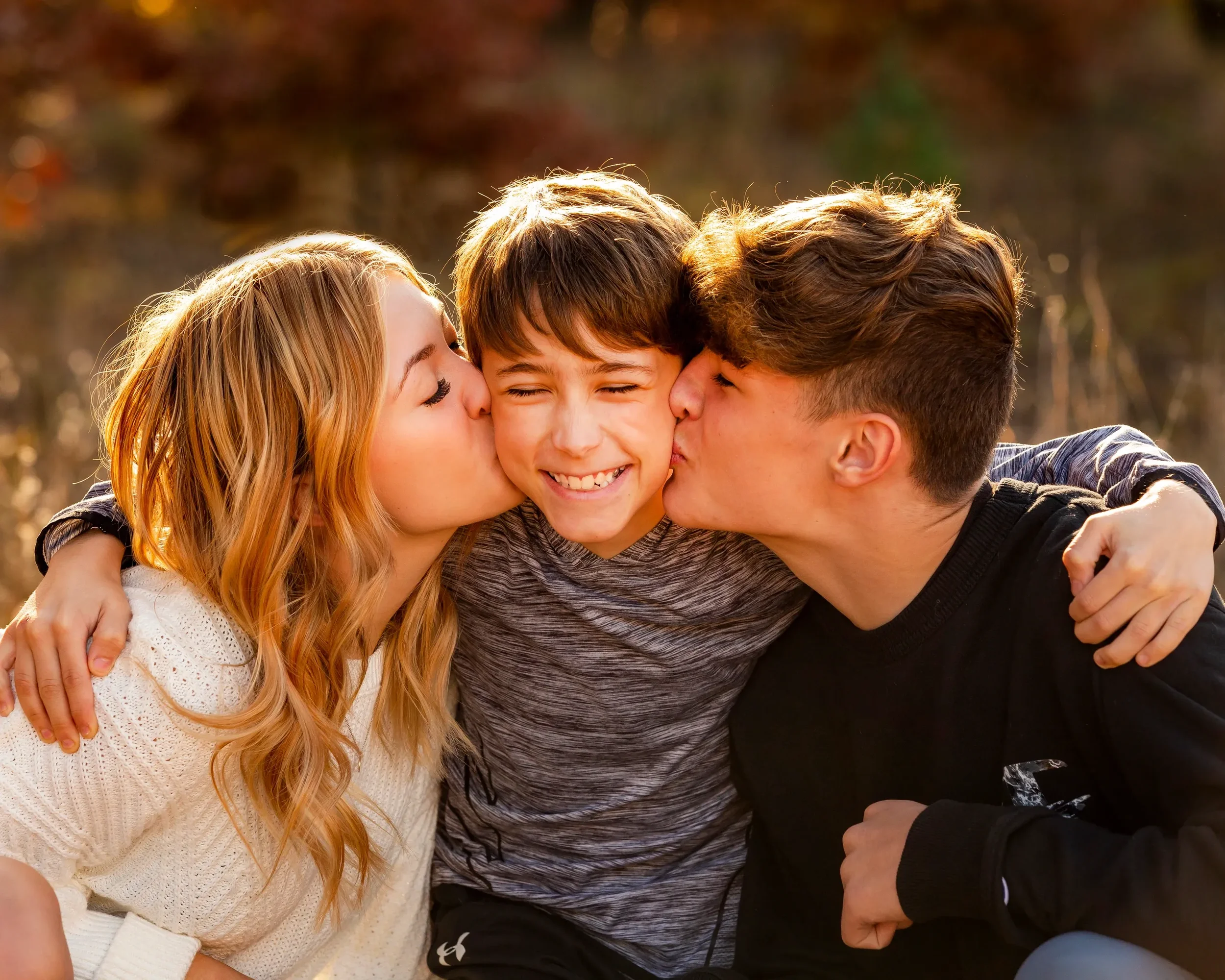 Three teenagers, two boys and a girl, hugging and kissing each other with smiles in an outdoor setting during fall.