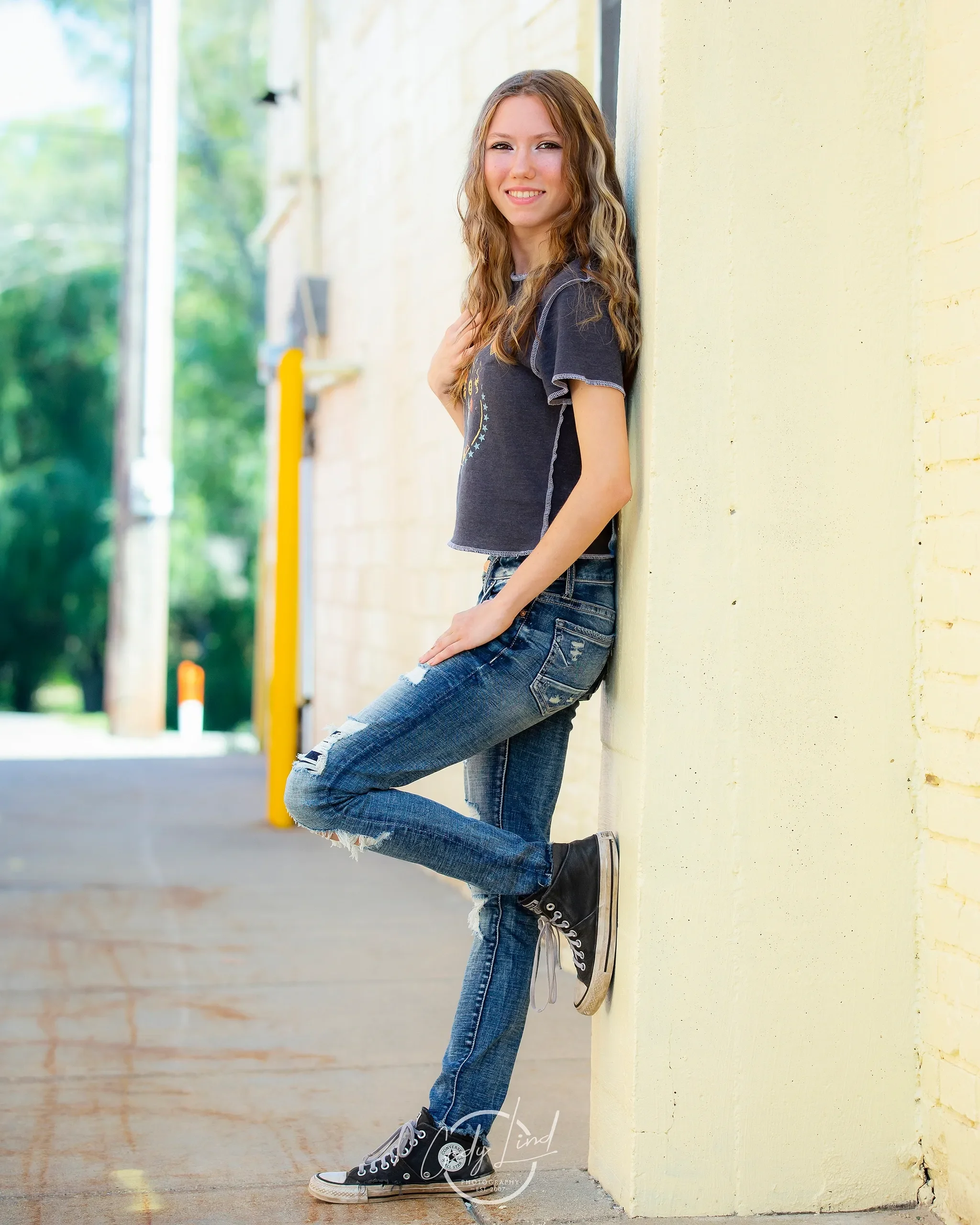 A young woman with long wavy hair is leaning against a yellow wall, smiling at the camera, wearing a black t-shirt, distressed jeans, and black sneakers.