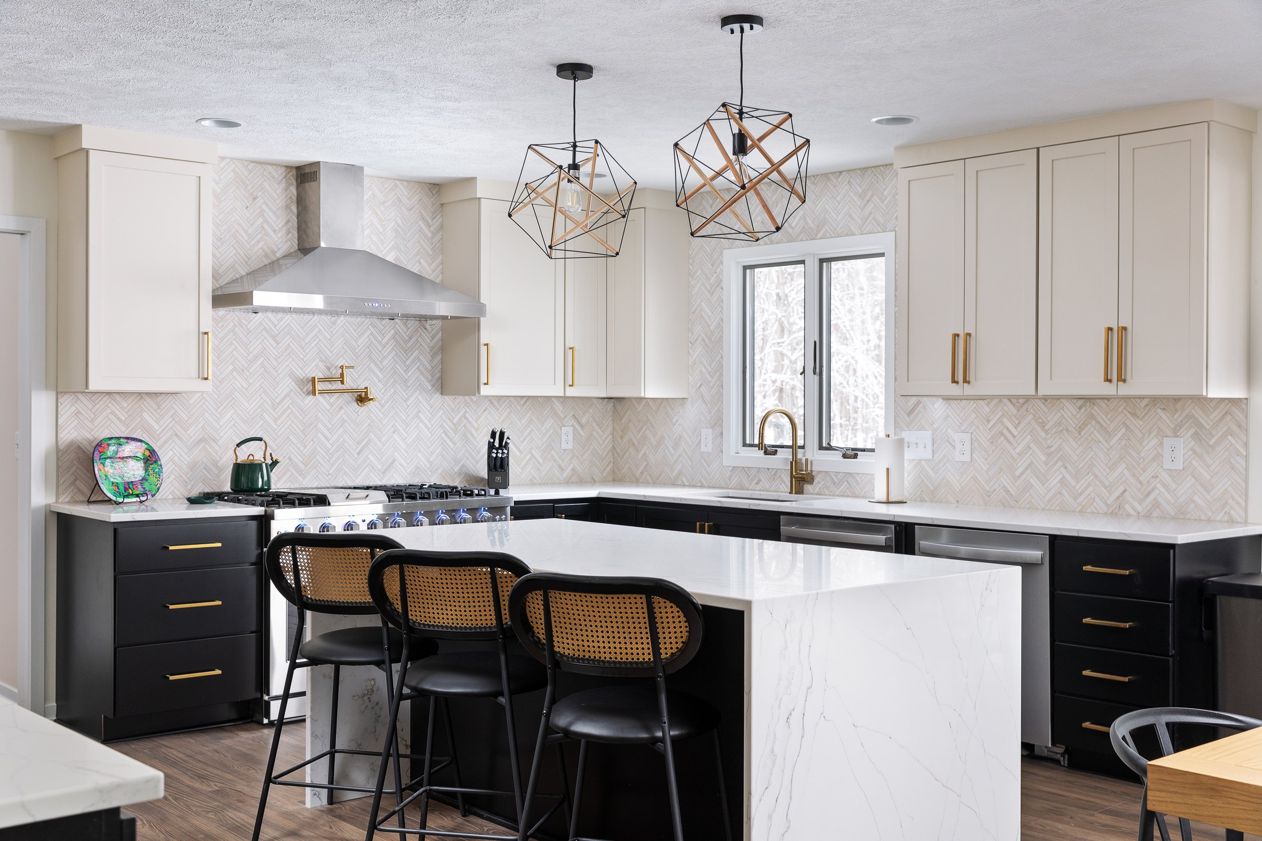 Modern kitchen with white cabinets, black lower cabinets with gold handles, a white marble island, and pendant lights with geometric frames. Stainless steel range hood, black countertops, and window showing snow outside.