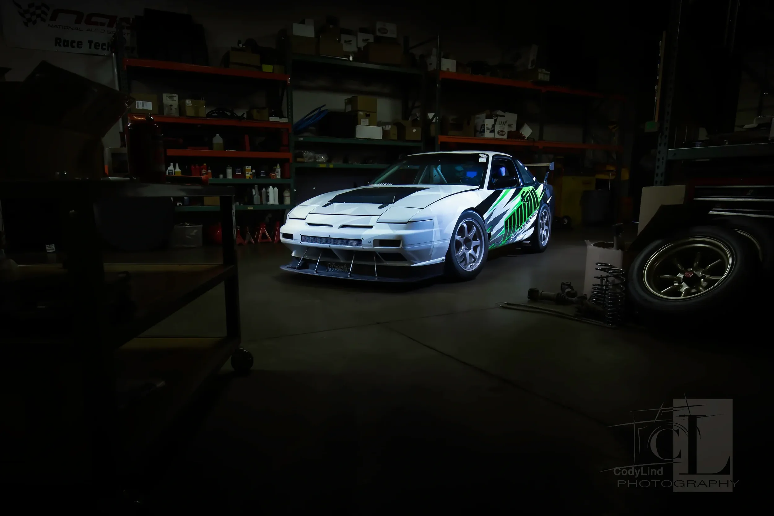 A white racing car with black, green, and gray graphics in a dimly lit garage, surrounded by shelves with tools and boxes, and some car parts on the floor.