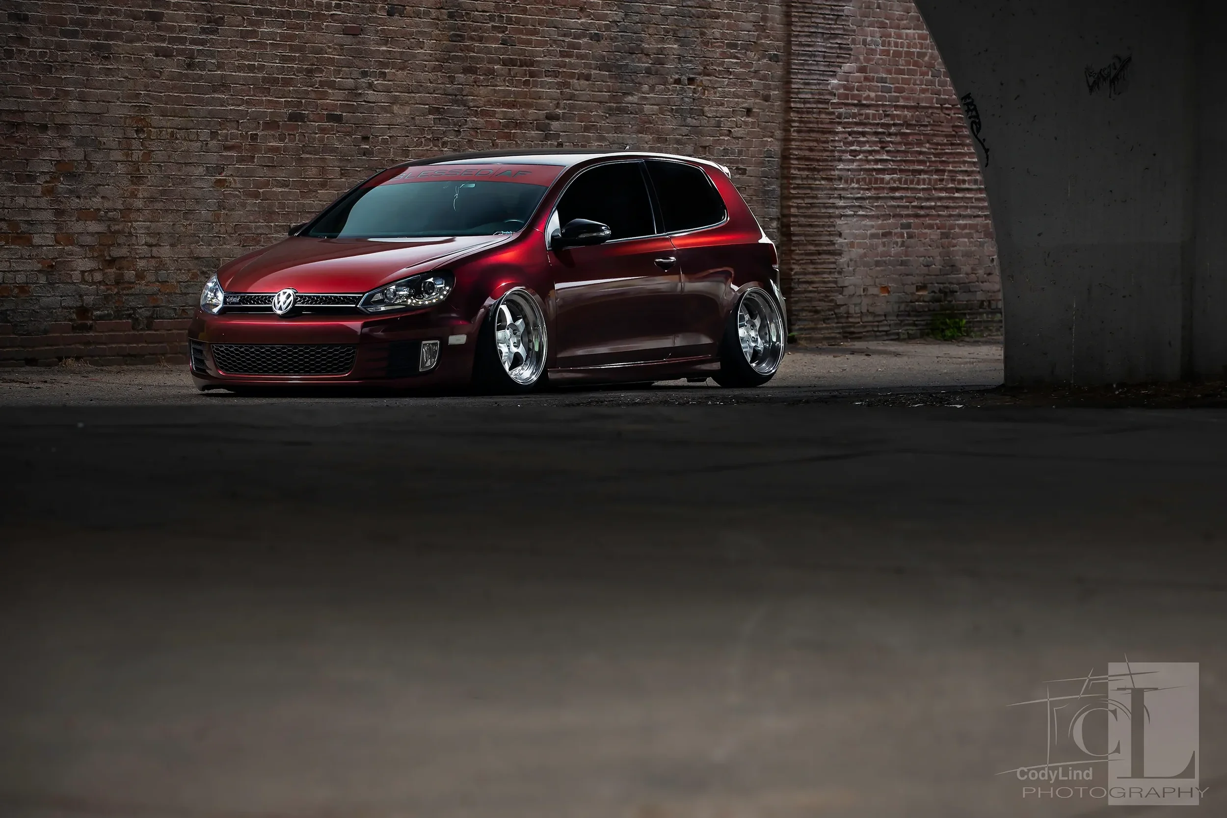 A lowered red Volkswagen hatchback car with chrome wheels parked against a brick wall in a dimly lit urban environment.