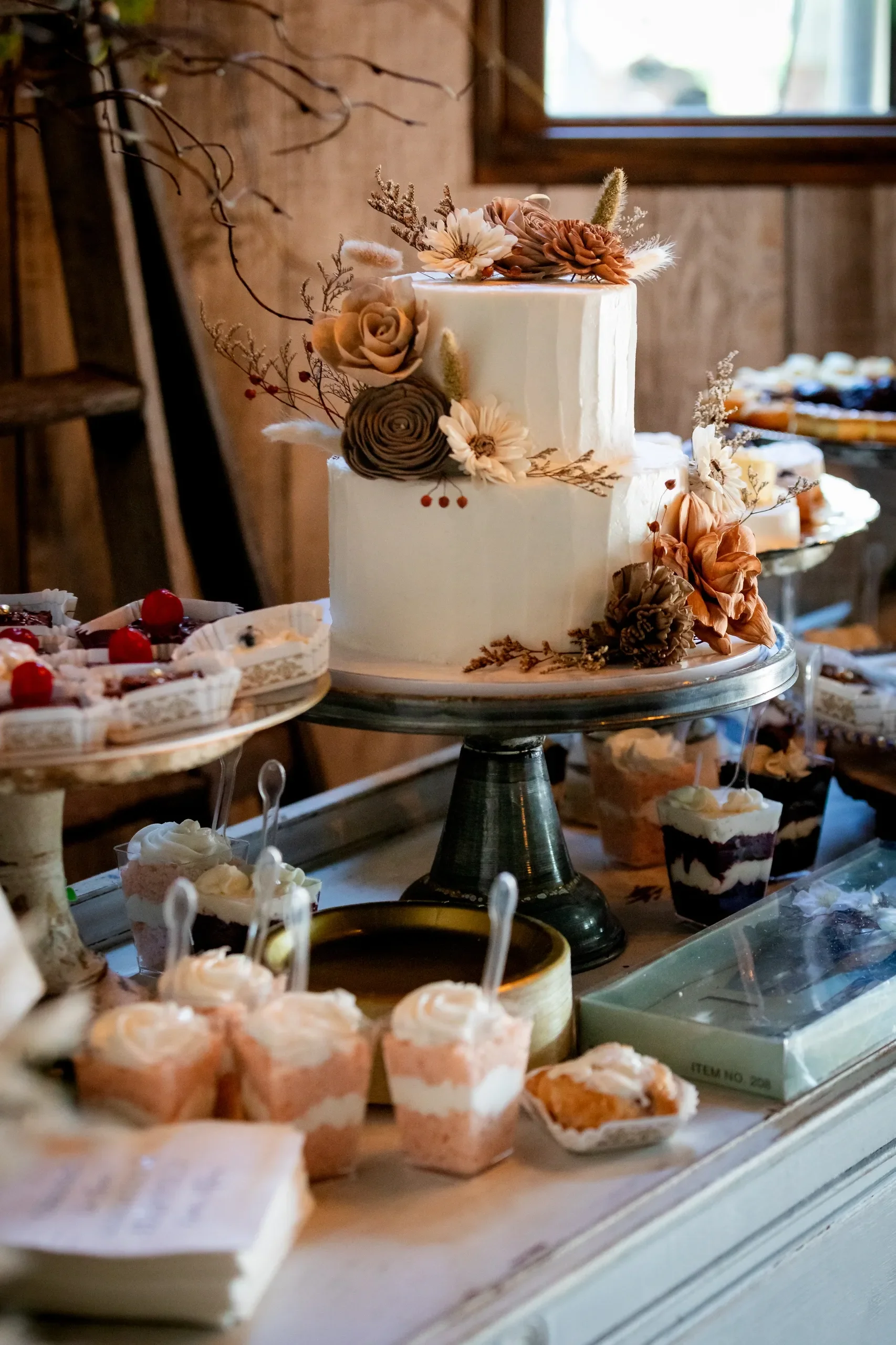 A white, two-tiered wedding cake decorated with dried flowers and foliage, set on a silver cake stand. Surrounding the cake are various small desserts including mini cupcakes, layered mousse cups, and pastries, displayed on a rustic wooden table in a