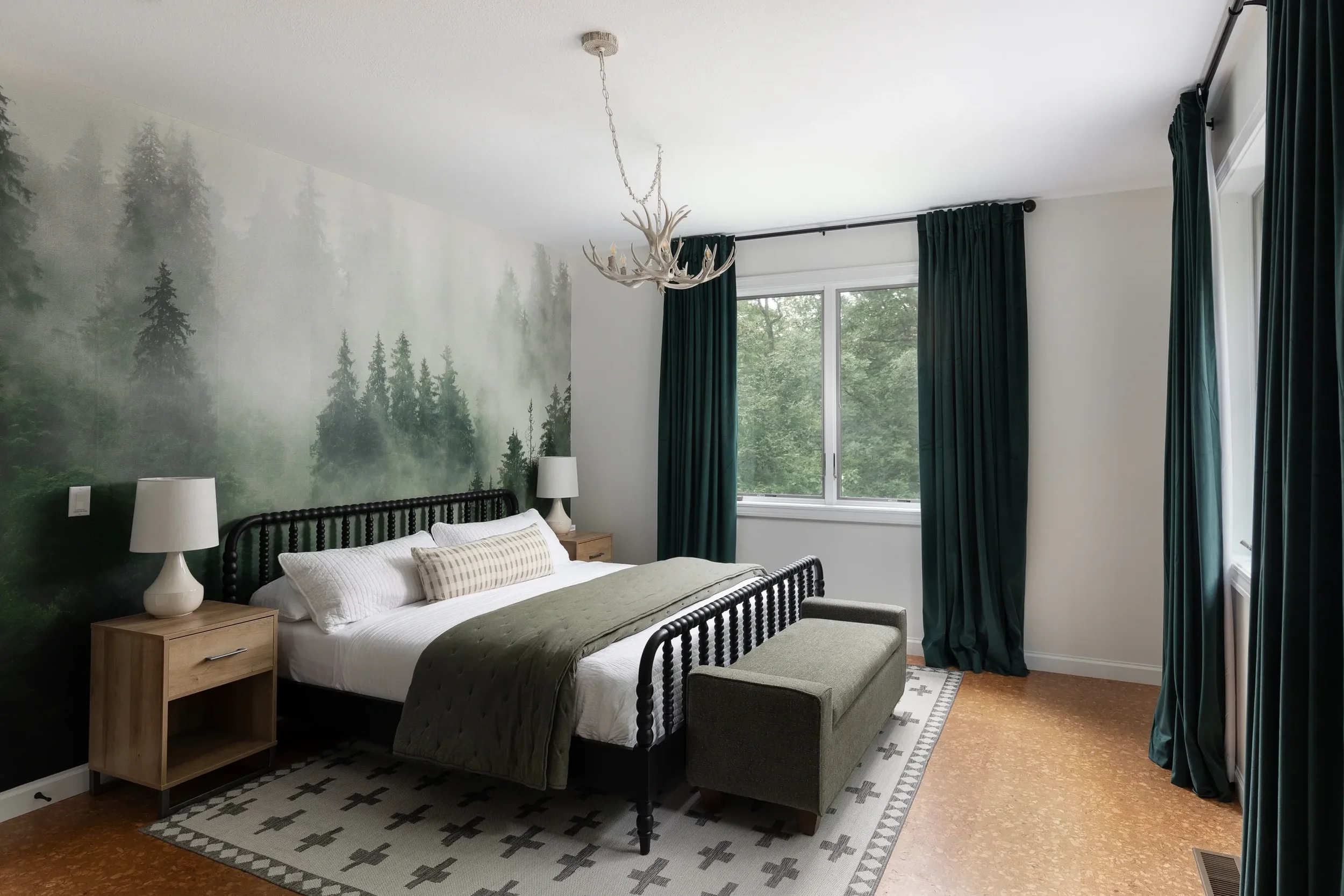 A bedroom with a large bed, dark wooden headboard, white bedding, and pillows. Two wooden nightstands with white lamps, a bench at the foot of the bed, a window with dark green curtains, and a mural of a forest scene on the wall behind the bed.
