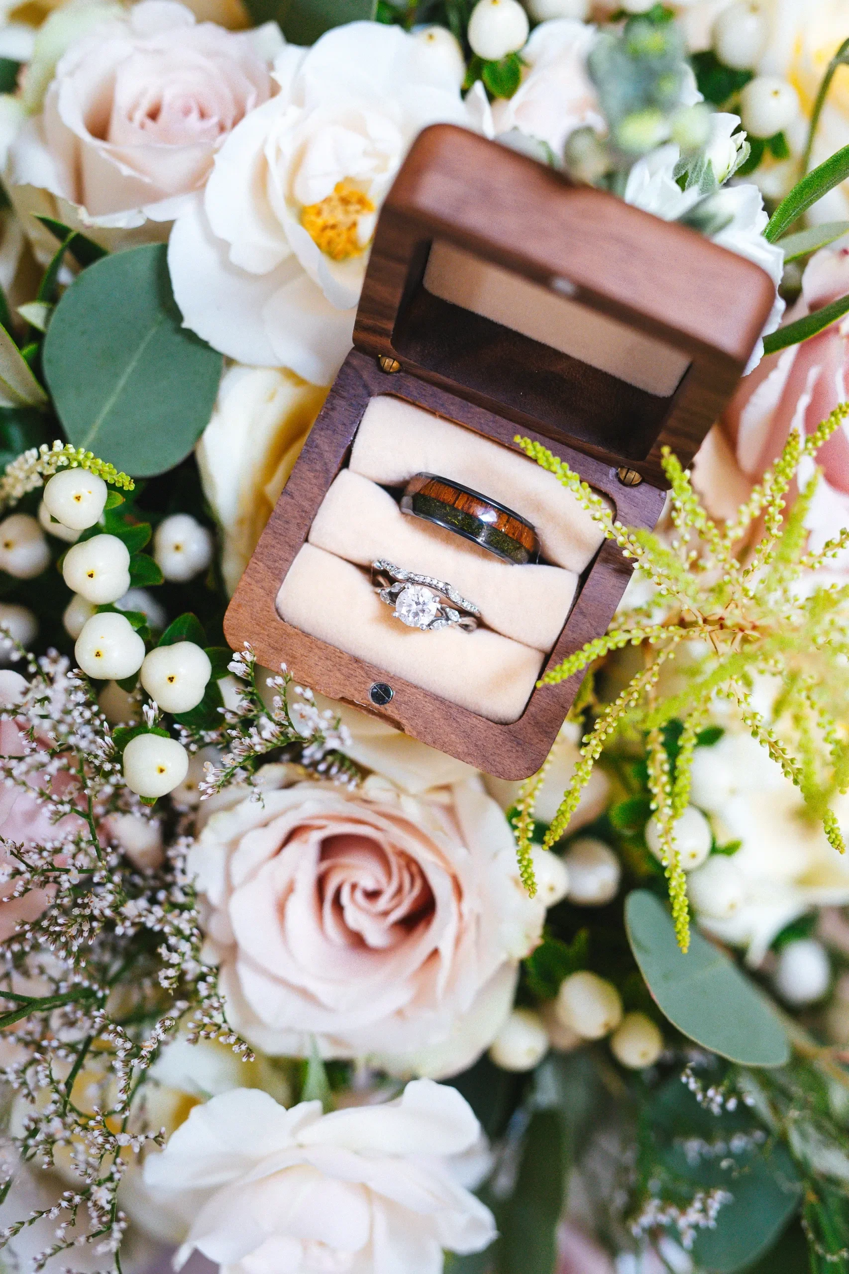 A wedding ring set in a wooden box surrounded by white and pink roses, berries, and greenery.