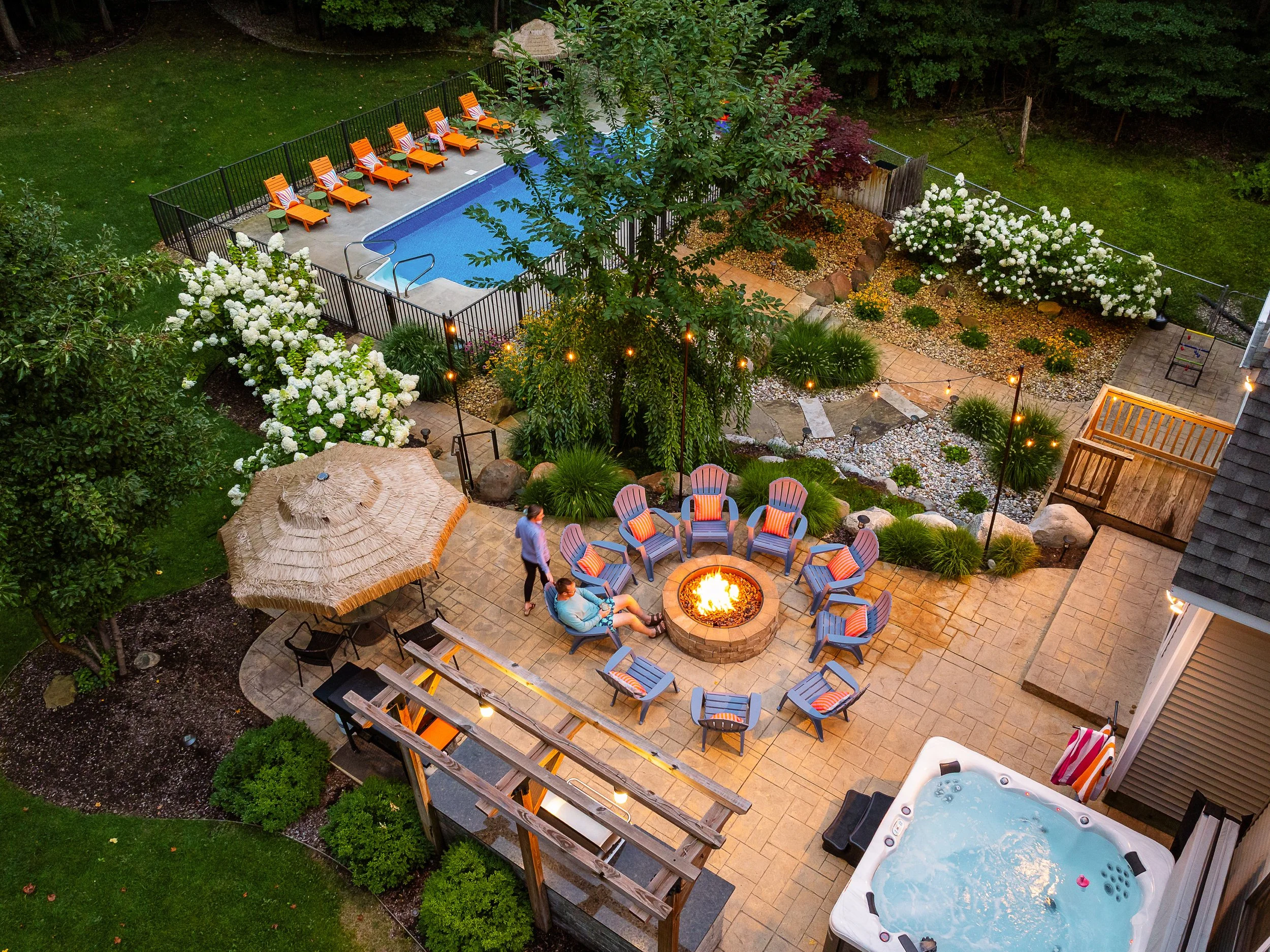 An aerial view of a backyard with a swimming pool, a patio area with a fire pit surrounded by chairs, a hot tub, and a landscaped garden with trees, white flowers, and string lights.