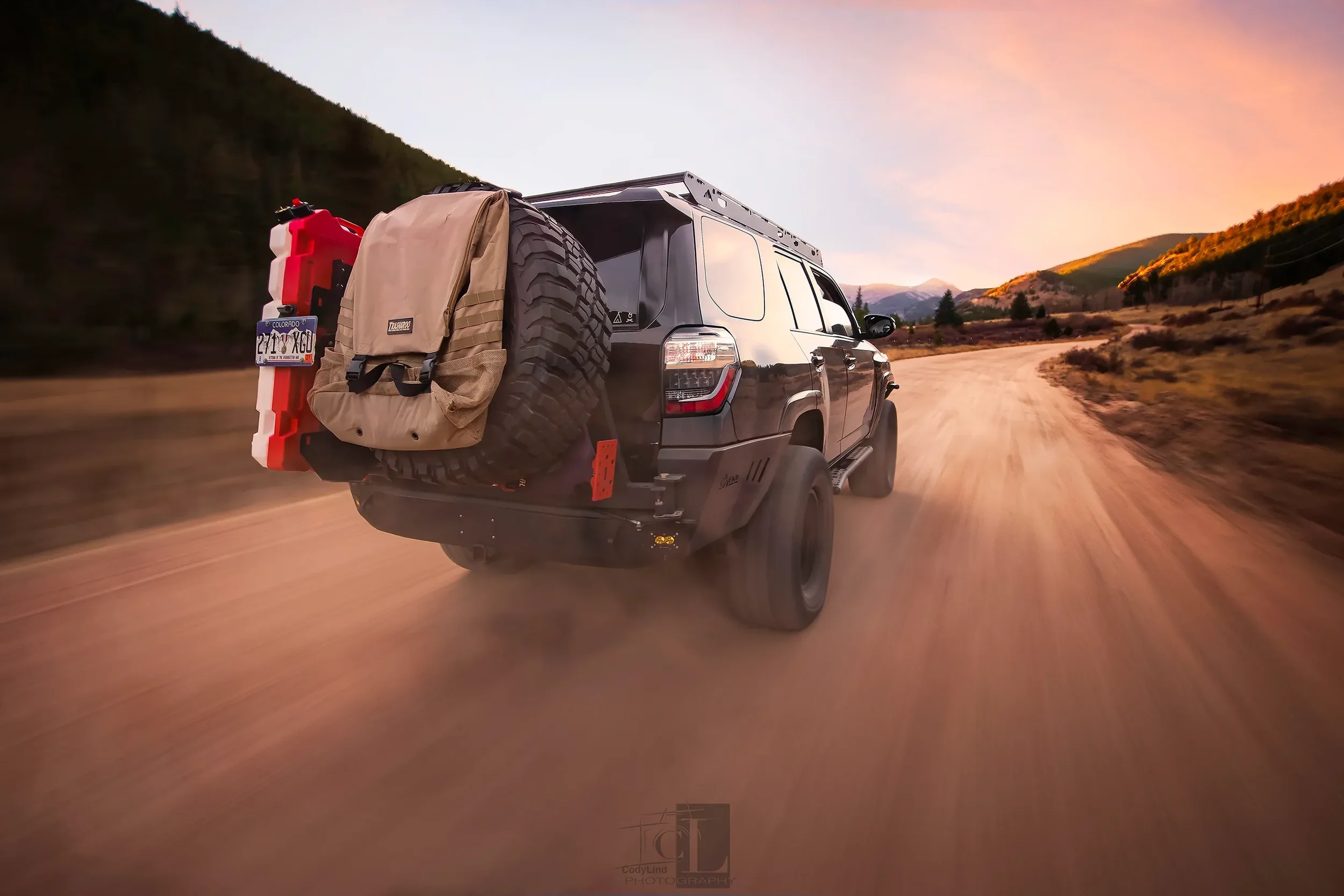 A black off-road SUV driving on a dirt road through a mountainous landscape during sunset.