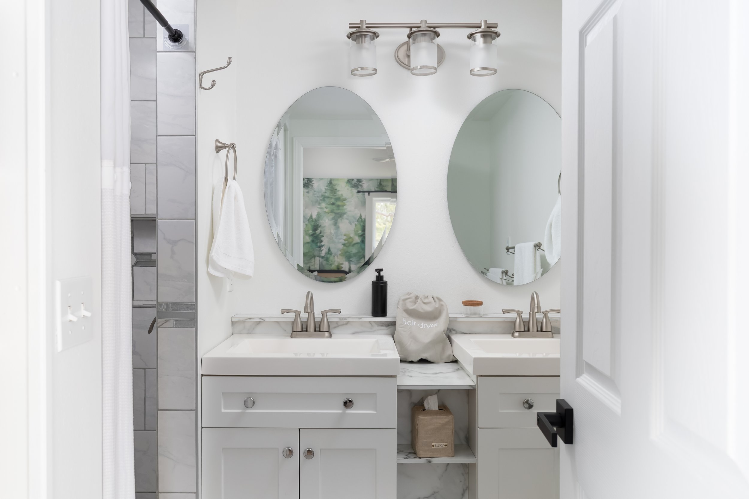 A bright bathroom with dual sinks, two oval mirrors, and a marble countertop. There is a black soap dispenser, a beige bag labeled 'hair dryer', and a tissue box beneath the counter. A towel hangs on a hook, and a wall-mounted light fixture is above 