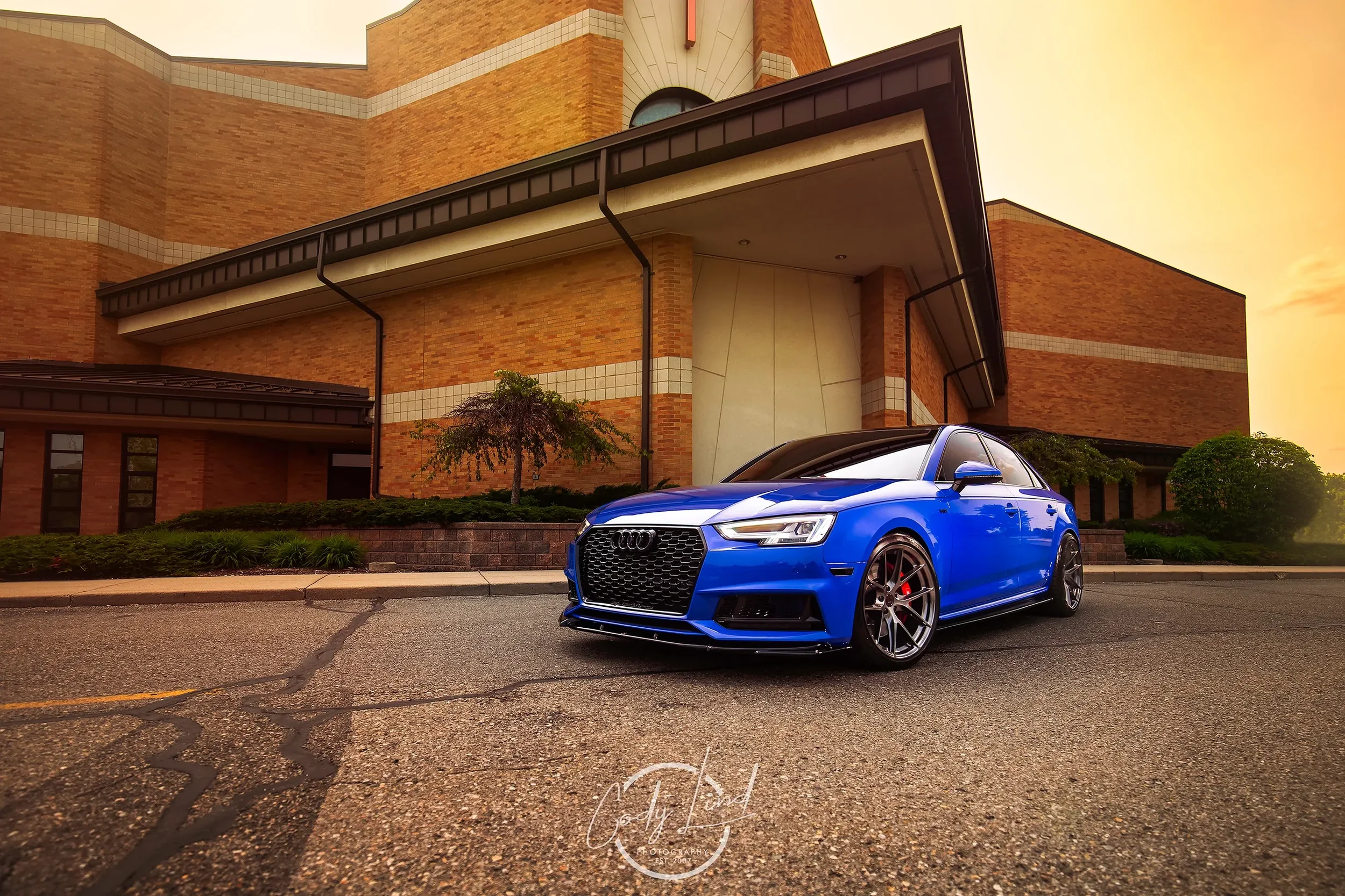 A blue Audi car parked on the street in front of a large brick building during sunset.