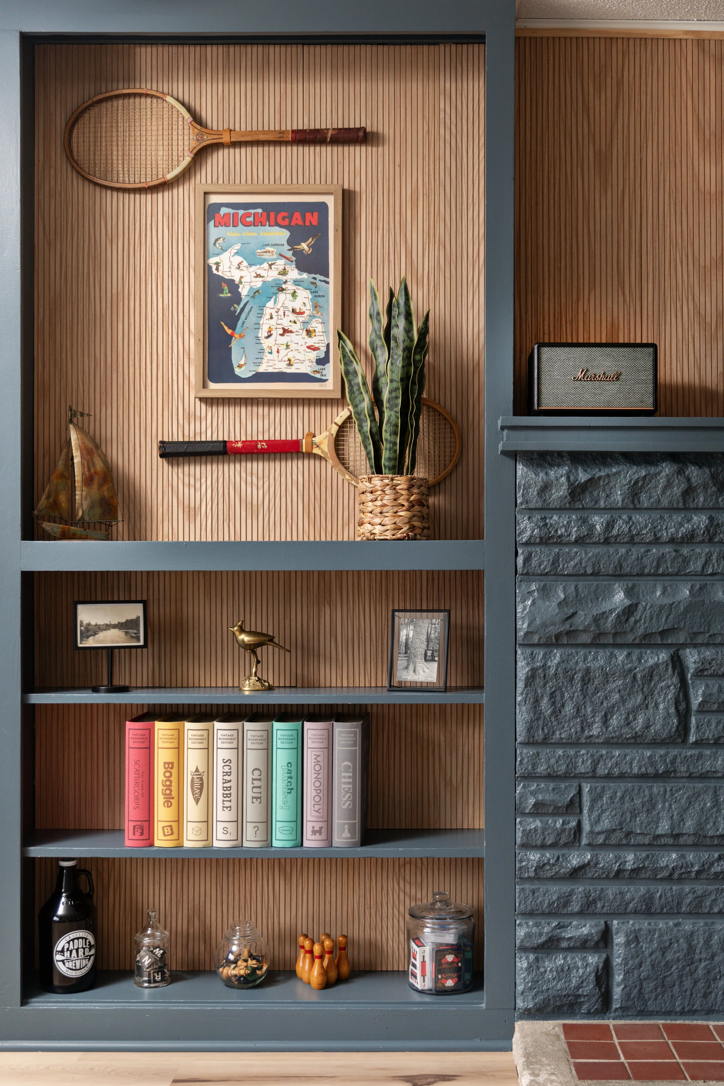A wooden bookshelf with collectibles including tennis rackets, a vintage paper sailboat, a plant in a woven basket, framed photos, a line of board game boxes, and miscellaneous jars and figurines. A Michigan map hangs on the wall with a racket above 