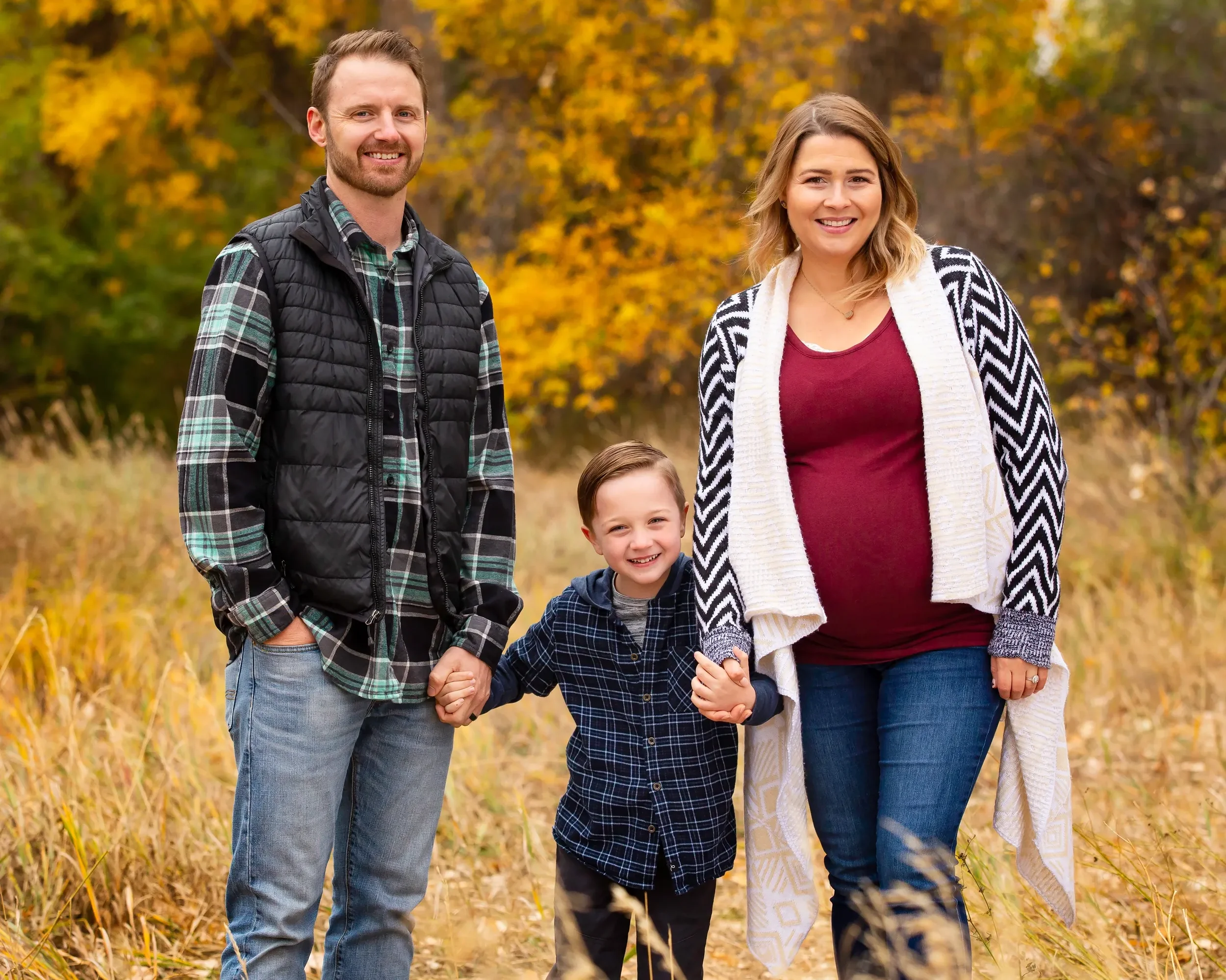 A family of three standing outdoors in a field during fall, smiling and holding hands. The father has a beard and wears a plaid shirt with a black vest, the mother has shoulder-length hair and wears a maroon top with a patterned cardigan, and the you