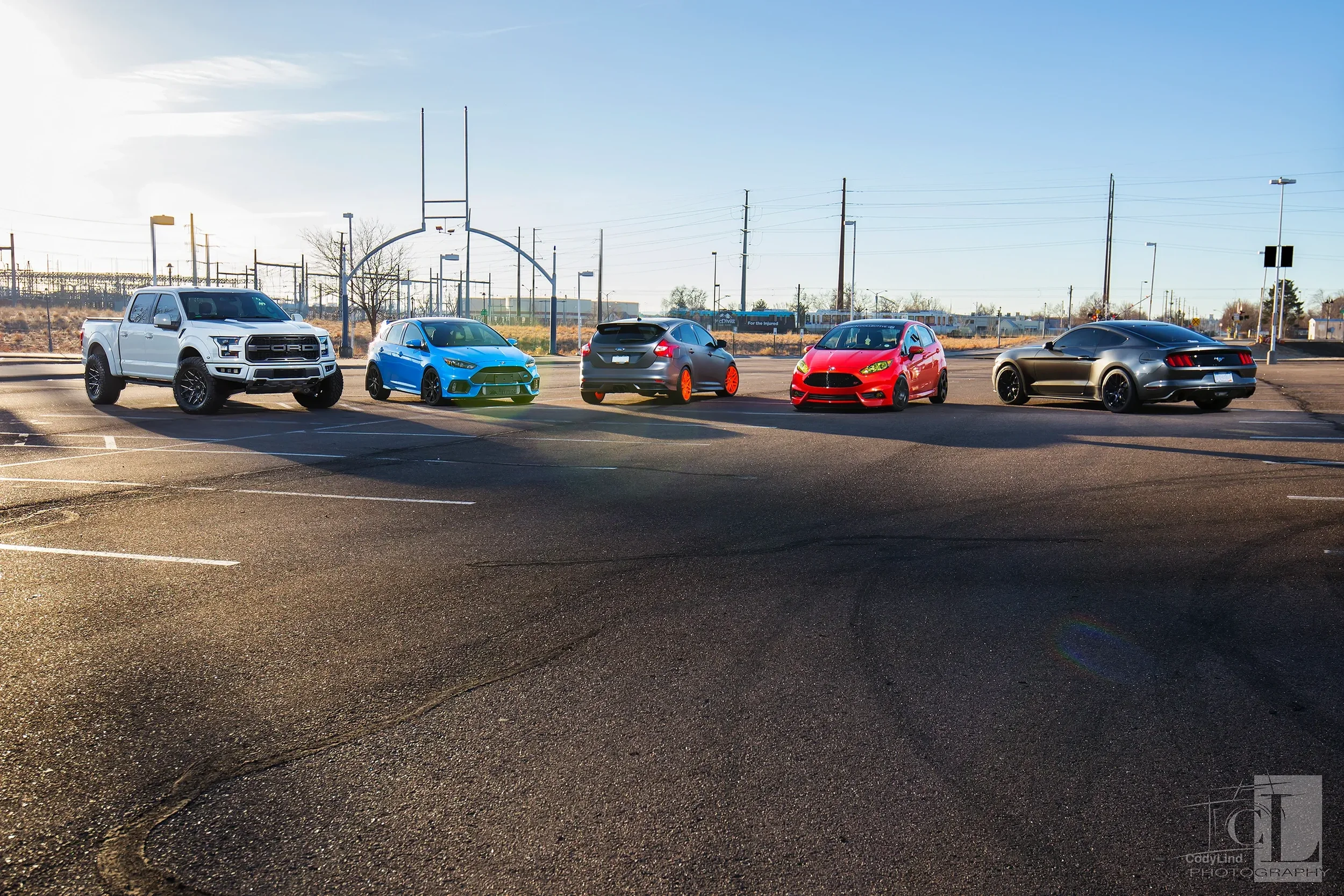 A parking lot with five parked cars of different colors, including white, blue, black, red, and black, with a football goalpost and power lines in the background and a clear sky.