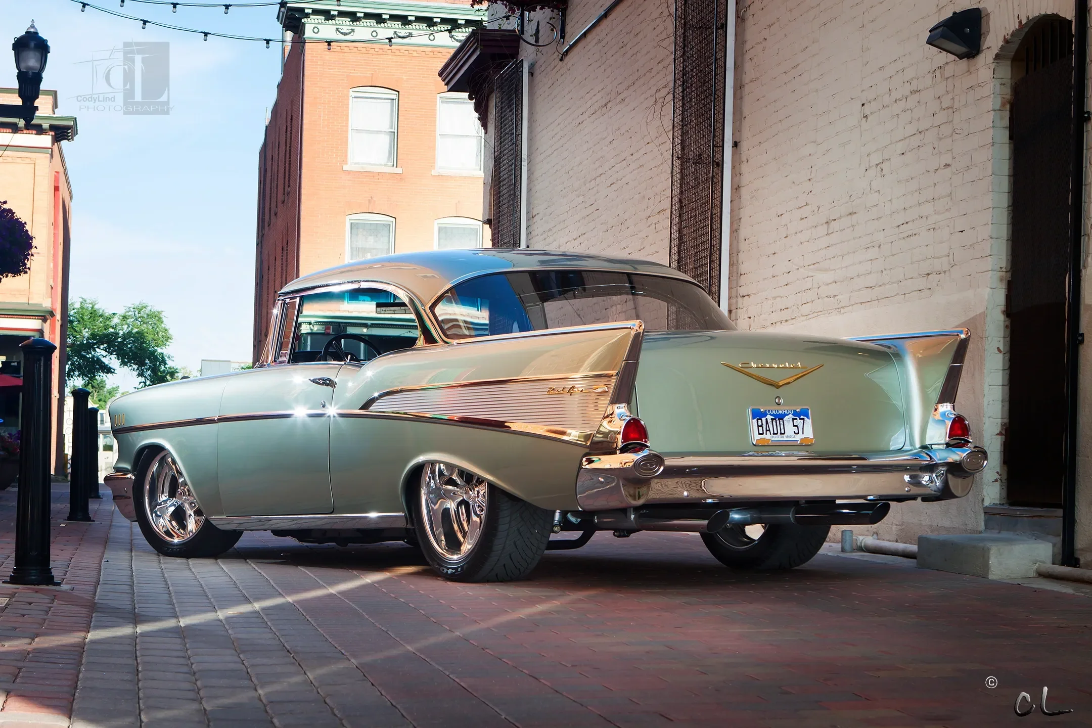 A vintage 1957 Chevrolet Bel Air parked on a brick sidewalk next to a brick building, with a blue Colorado license plate reading "BAUU 57." The car has a two-tone paint job, with the upper part in silver and the lower part in light green, featuring c