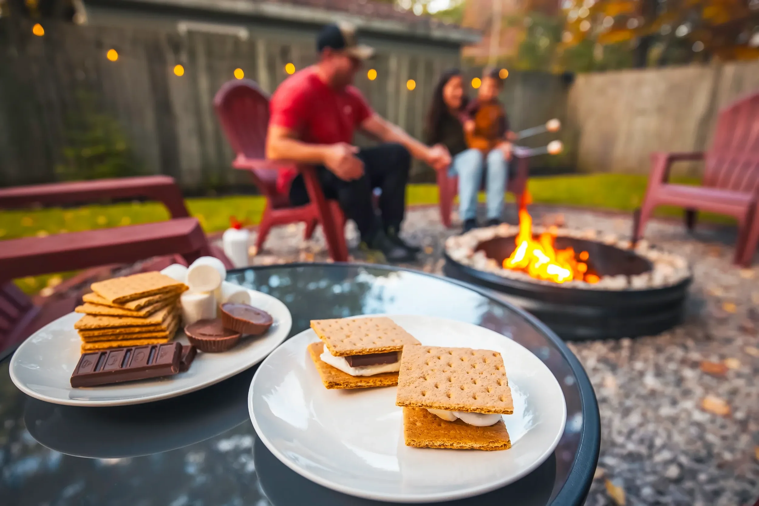 S'mores with graham crackers, marshmallows, and chocolate on a white plate in the foreground, with people sitting around a fire pit in the background in a backyard.