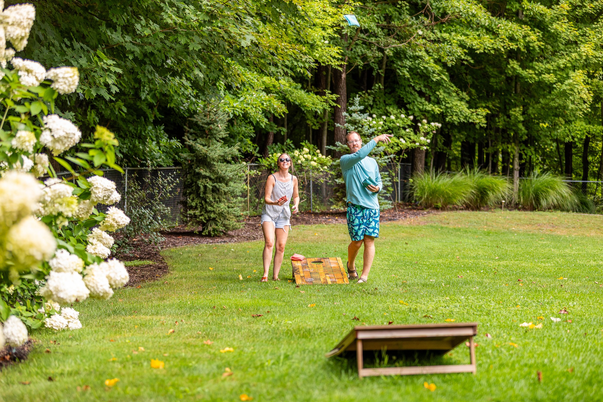A man and woman playing cornhole in a backyard with green grass, trees, and shrubs.