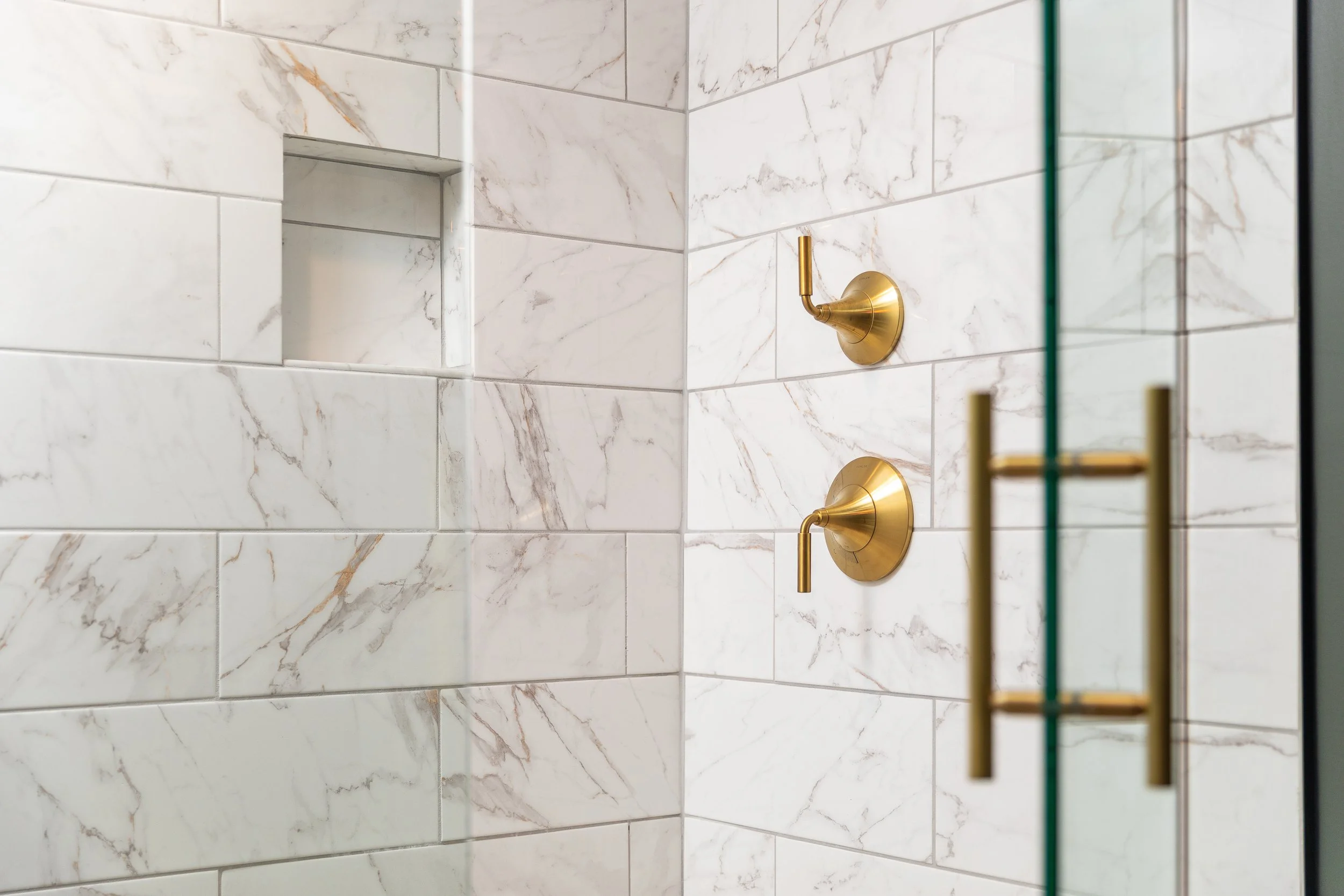 Close-up of a marble-tiled shower wall with two gold shower controls and a built-in shower niche.
