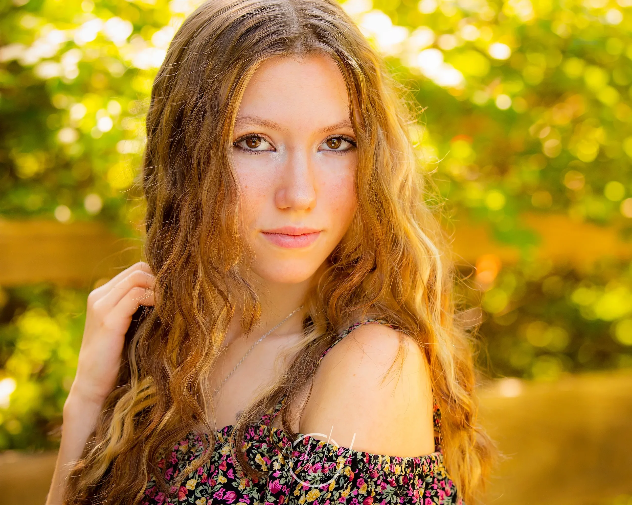 Portrait of a young woman with long, curly red hair and hazel eyes, wearing a floral off-shoulder top, outdoors with blurred greenery in the background.
