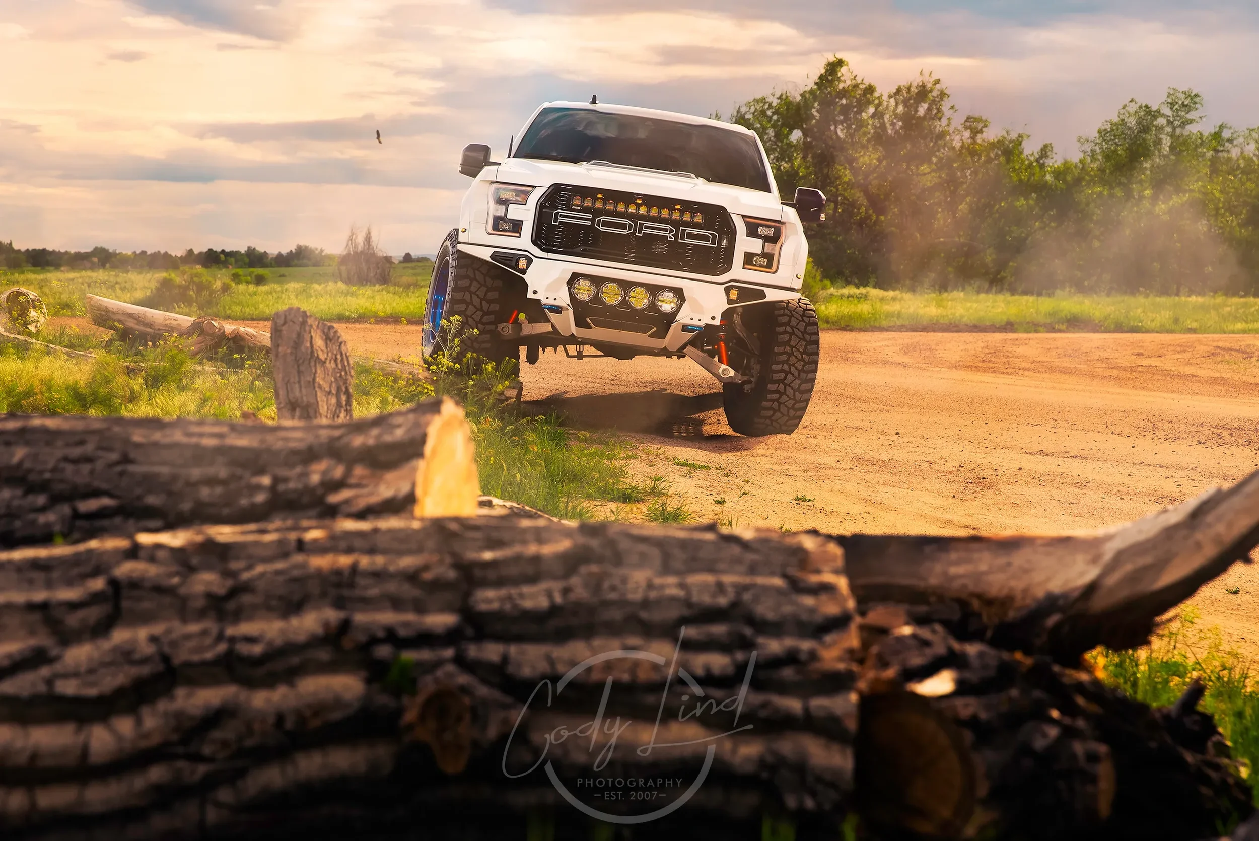 A white off-road truck with large tires driving on a dirt road in a countryside landscape, with logs in the foreground and green trees in the background during sunset.