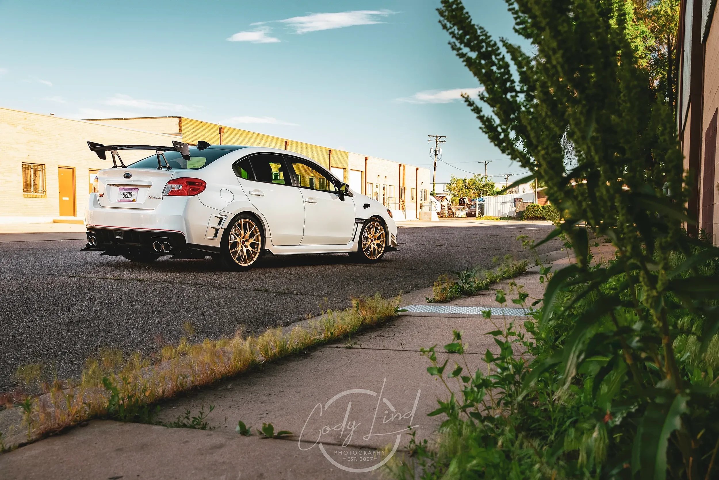 White sports sedan with a large rear spoiler, gold wheels, and quad exhaust pipes parked on an urban street scene during daytime.