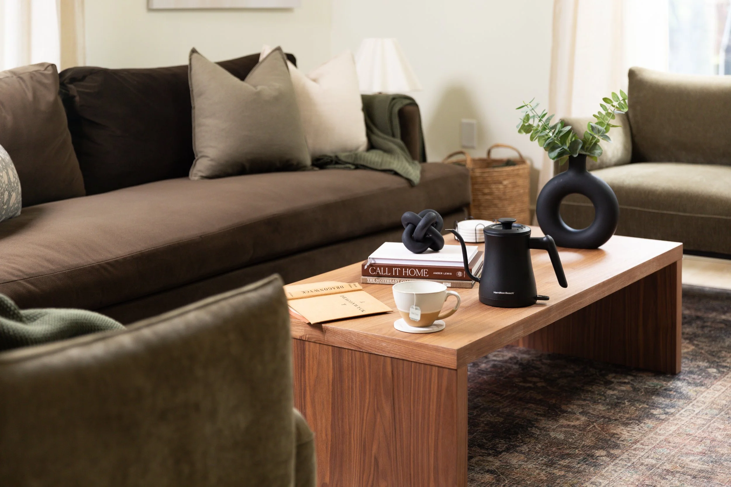 A living room with a brown sofa adorned with light-colored pillows, a wooden coffee table with a black teapot, a white mug, books, and decorative vases with greenery, and a woven basket in the background.