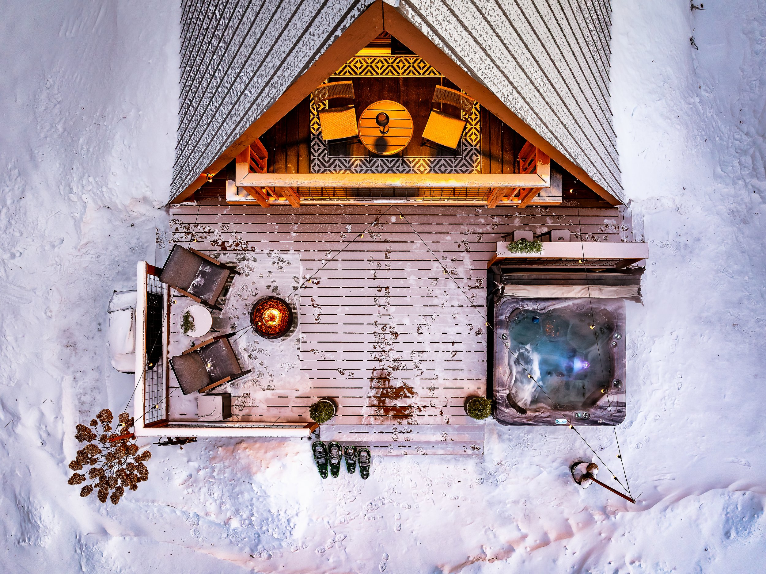 Top-down view of an outdoor snow-covered deck with a hot tub, patio furniture, a fire pit, and decorated with small potted plants, enclosed by snow-covered ground.