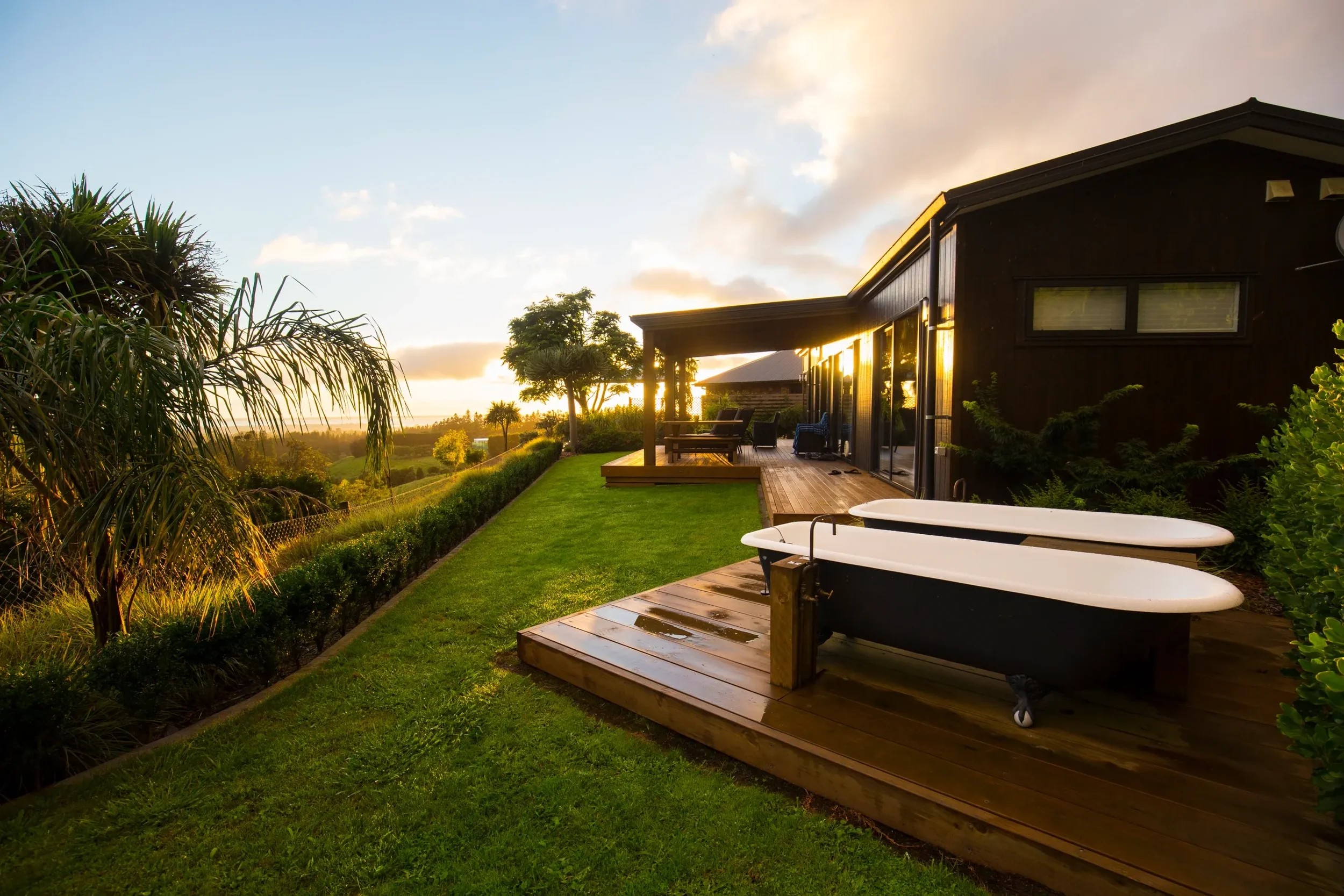 Sunset view of a backyard patio with two bathtubs on a wooden deck, adjacent to a black house with large windows, surrounded by greenery and palm trees.