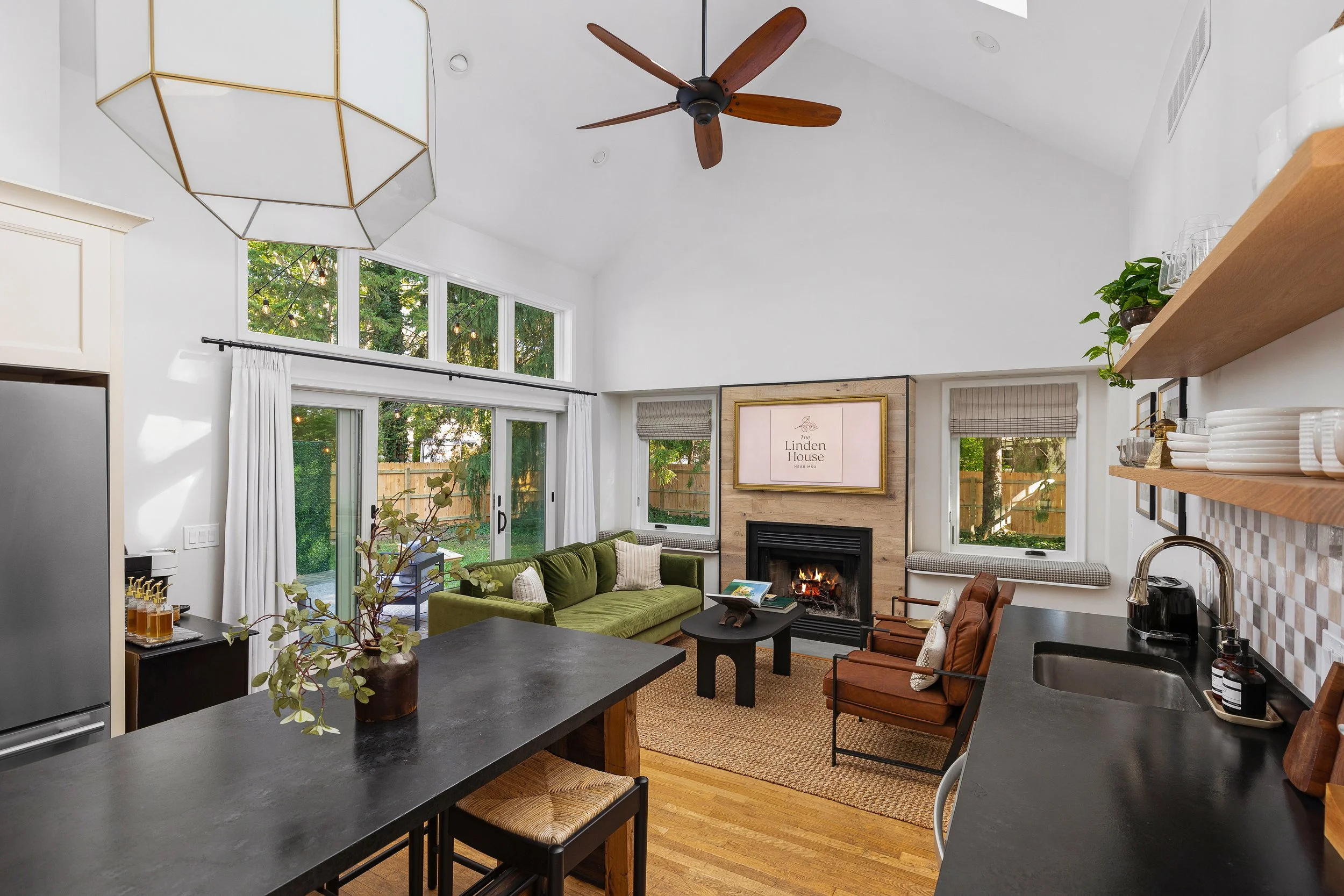 Living room with high ceiling, large windows, and a ceiling fan. Features a green sofa, armchairs, a fireplace, and wall-mounted TV. Open kitchen with black countertops and wooden shelves.