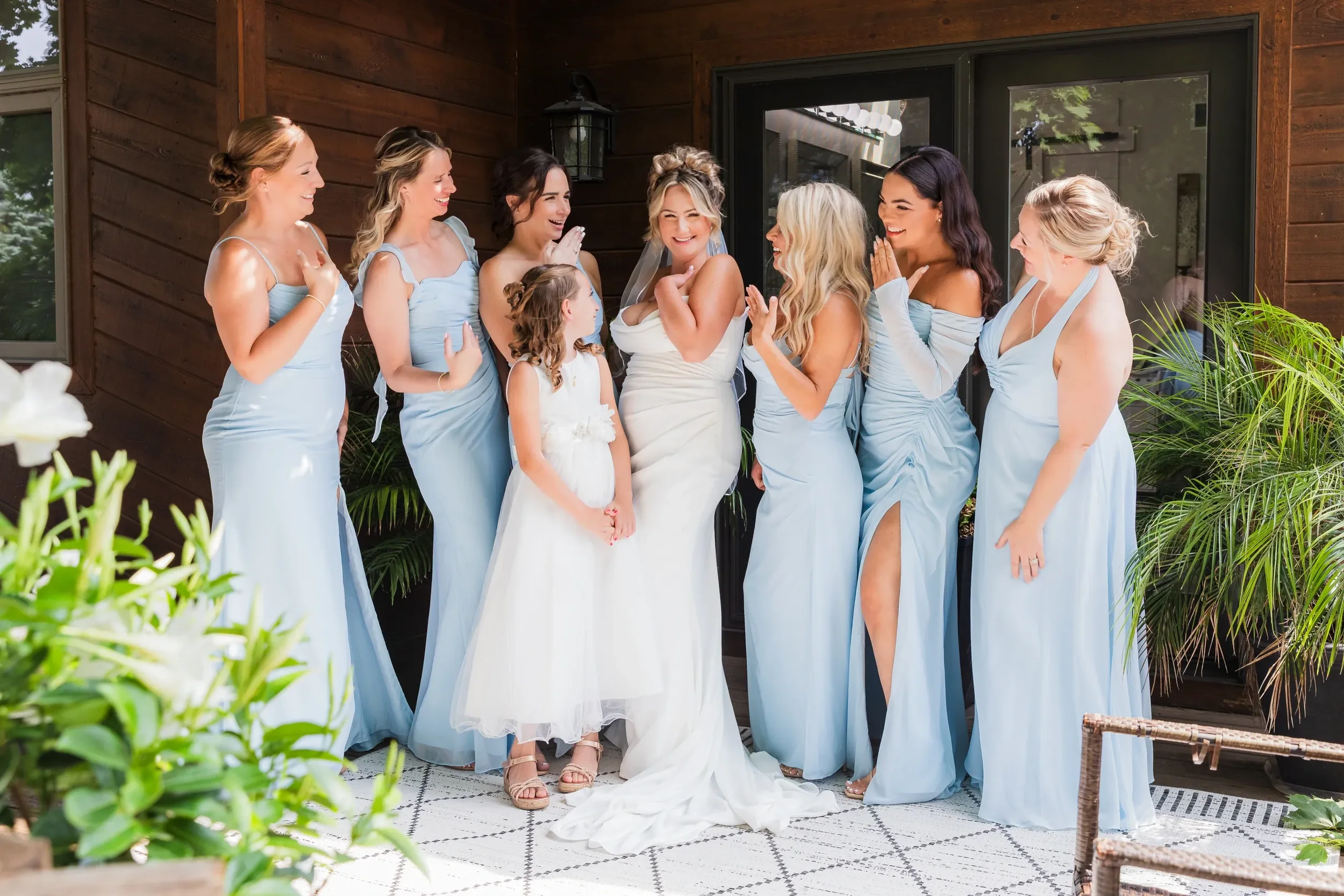 Bride with veil and white dress surrounded by smiling bridesmaids in light blue dresses. One young girl in white dress also present. They are outside on a porch with wood siding and green plants, celebrating with laughter.