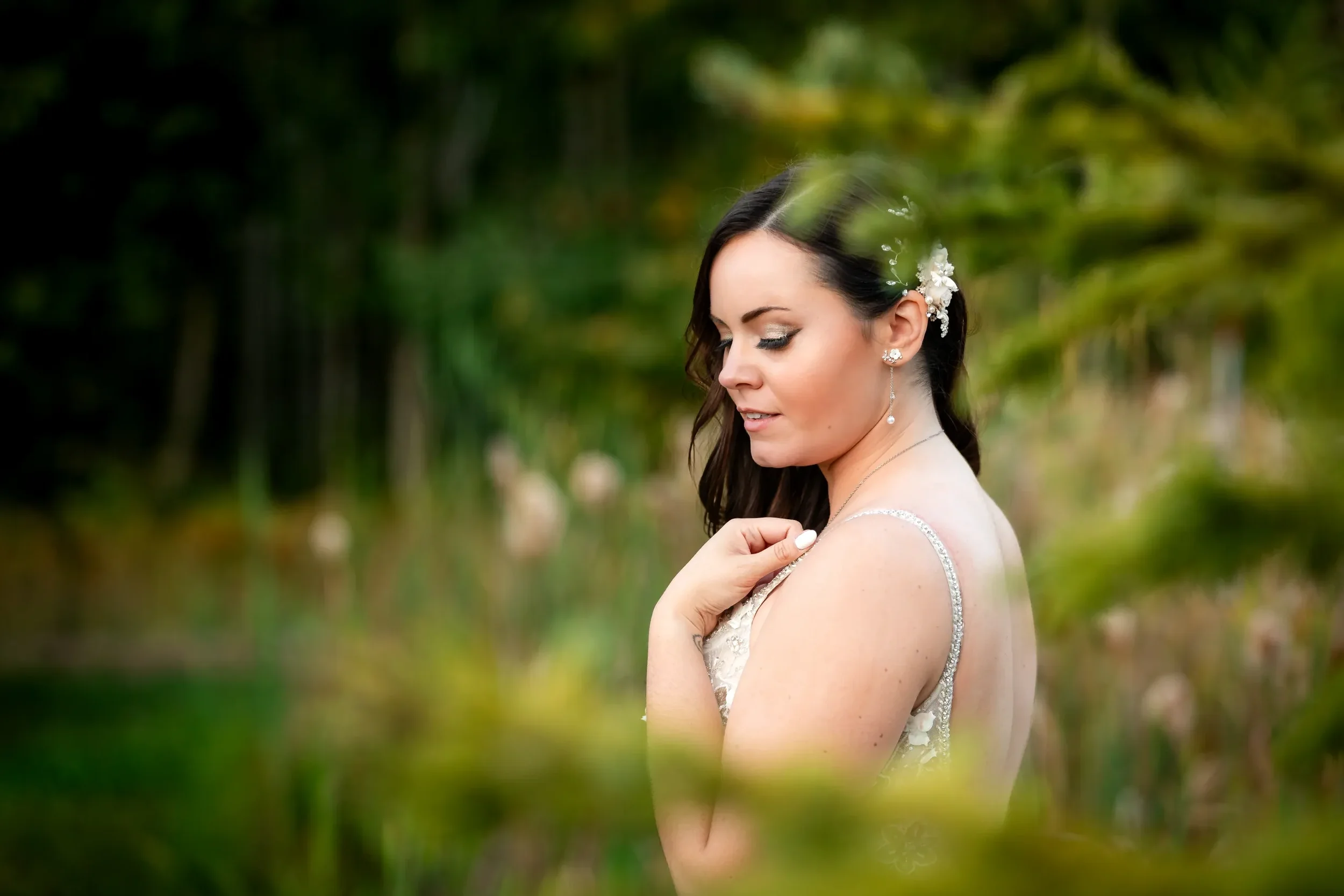 A bride with dark hair and a floral hairpiece, dressed in a white wedding gown, standing outdoors among greenery, looking down with her hand on her shoulder.