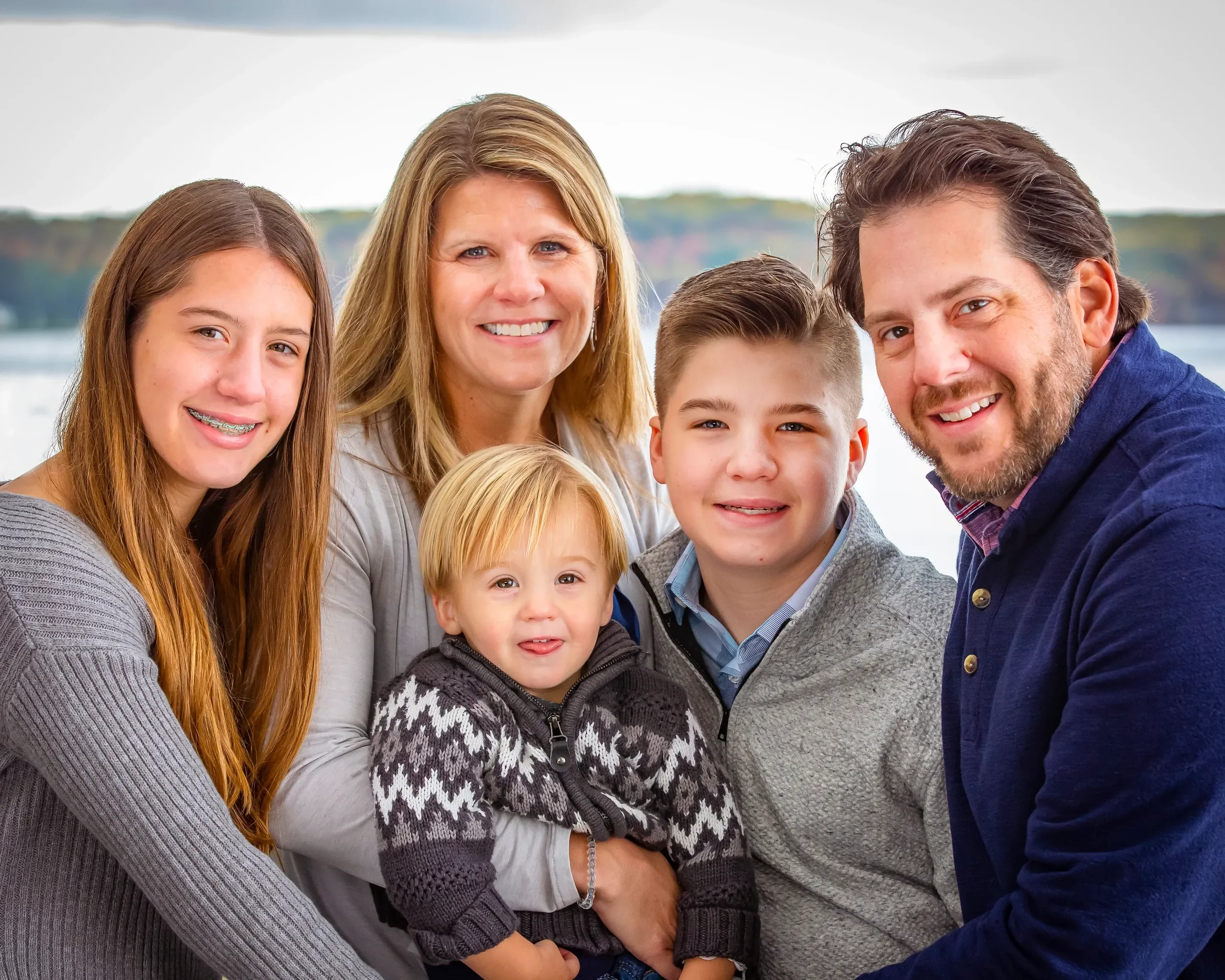 Family of six posing outdoors near a body of water, smiling for the camera, with trees in the background.