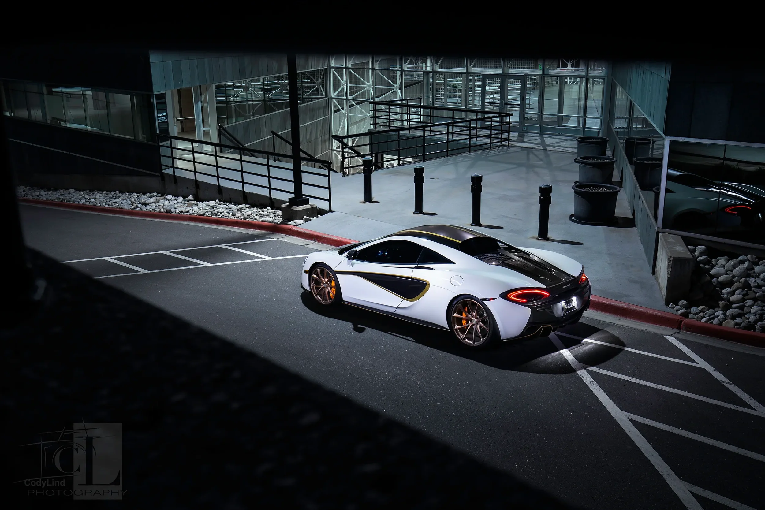 A white and black sports car with gold rims parked in a dimly lit parking lot at night, near a modern building entrance. mclaren 