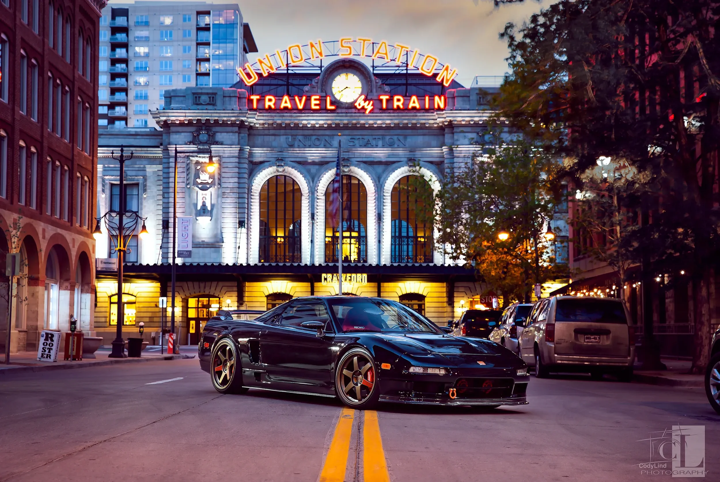 A black sports car parked on a city street in front of Union Station during twilight, with vintage-style street lamps, city buildings, and trees illuminated in the background.