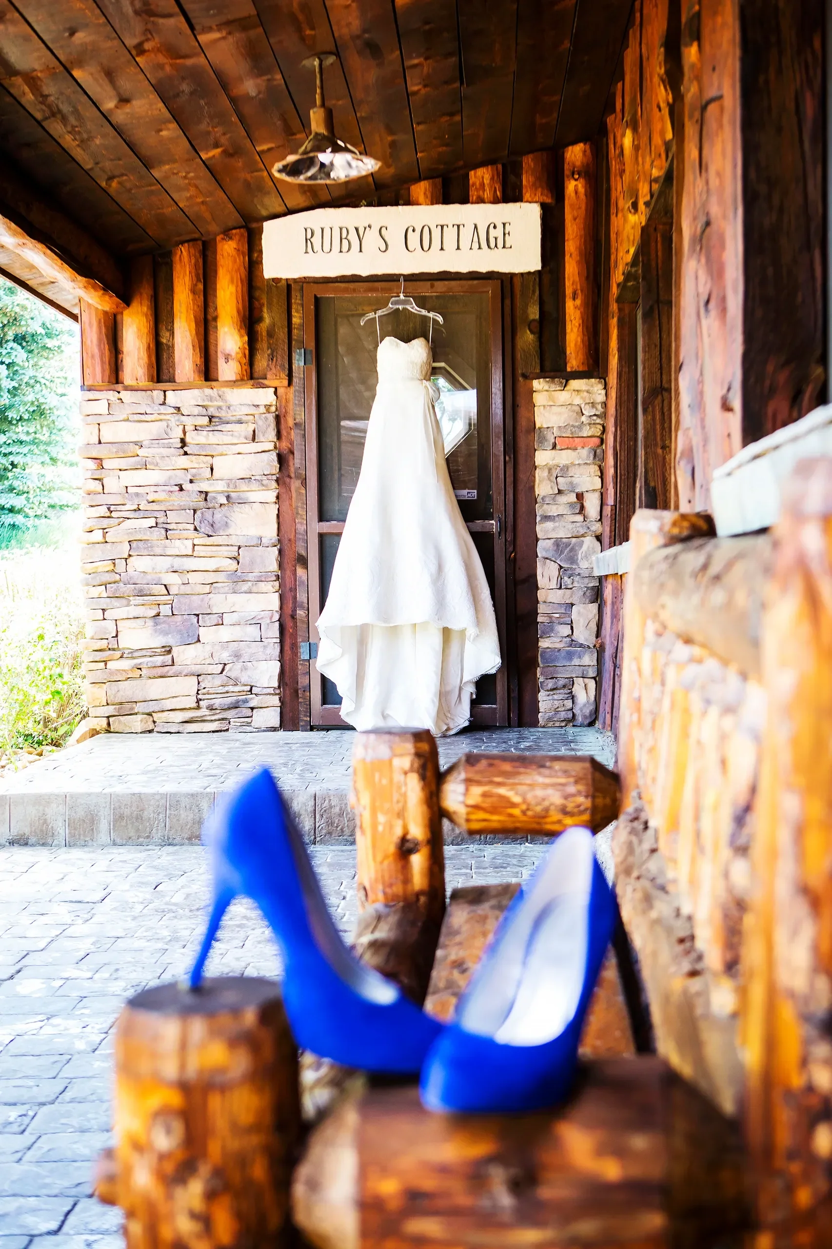 A wedding dress hanging on a hanger in front of a window with a sign that says 'Ruby's Cottage'; a pair of bright blue high heels on a wooden bench in a rustic, wood and stone porch setting.