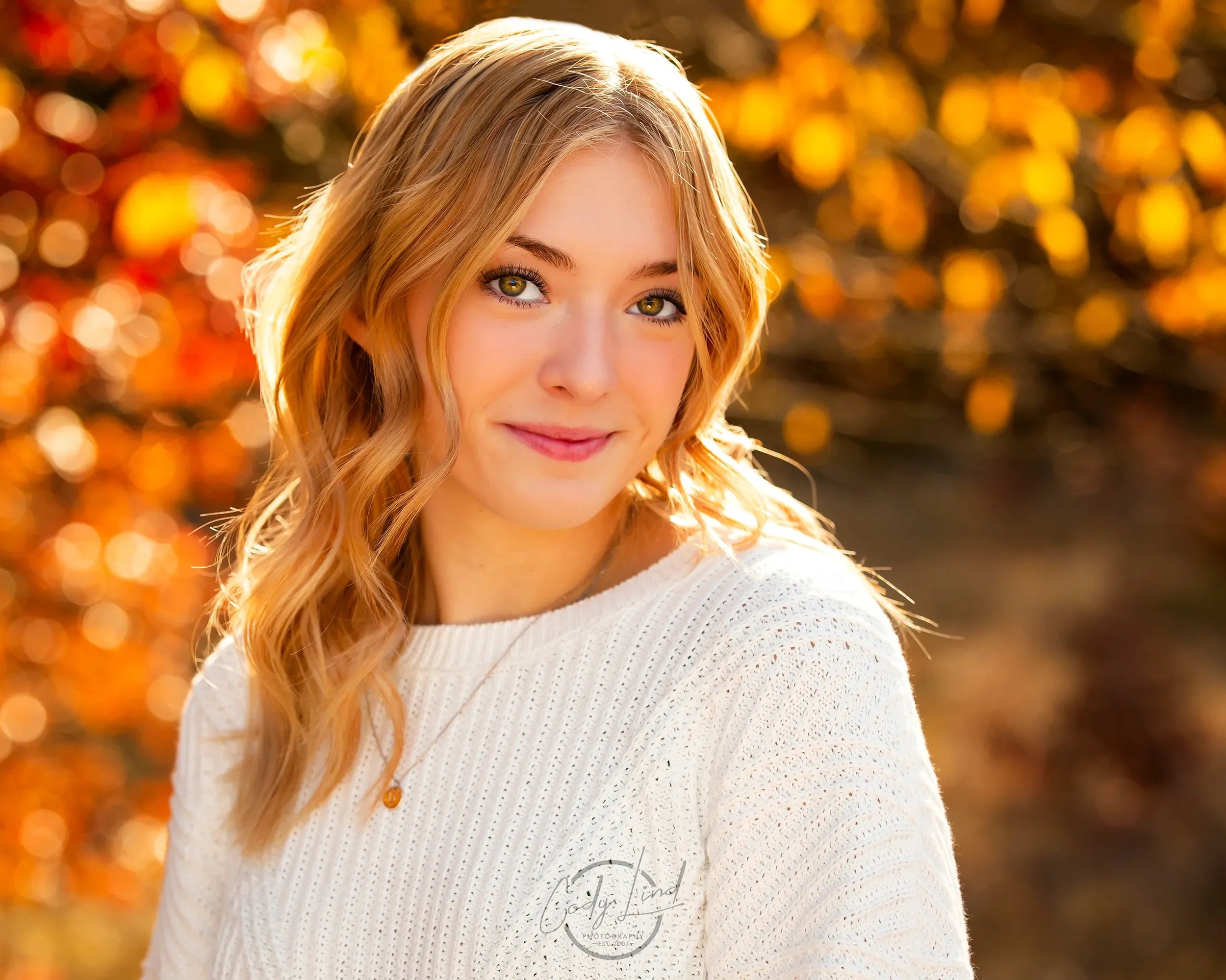 A young woman with wavy blonde hair and green eyes smiling outdoors during fall, wearing a white knit sweater, with orange and yellow autumn leaves in the background.