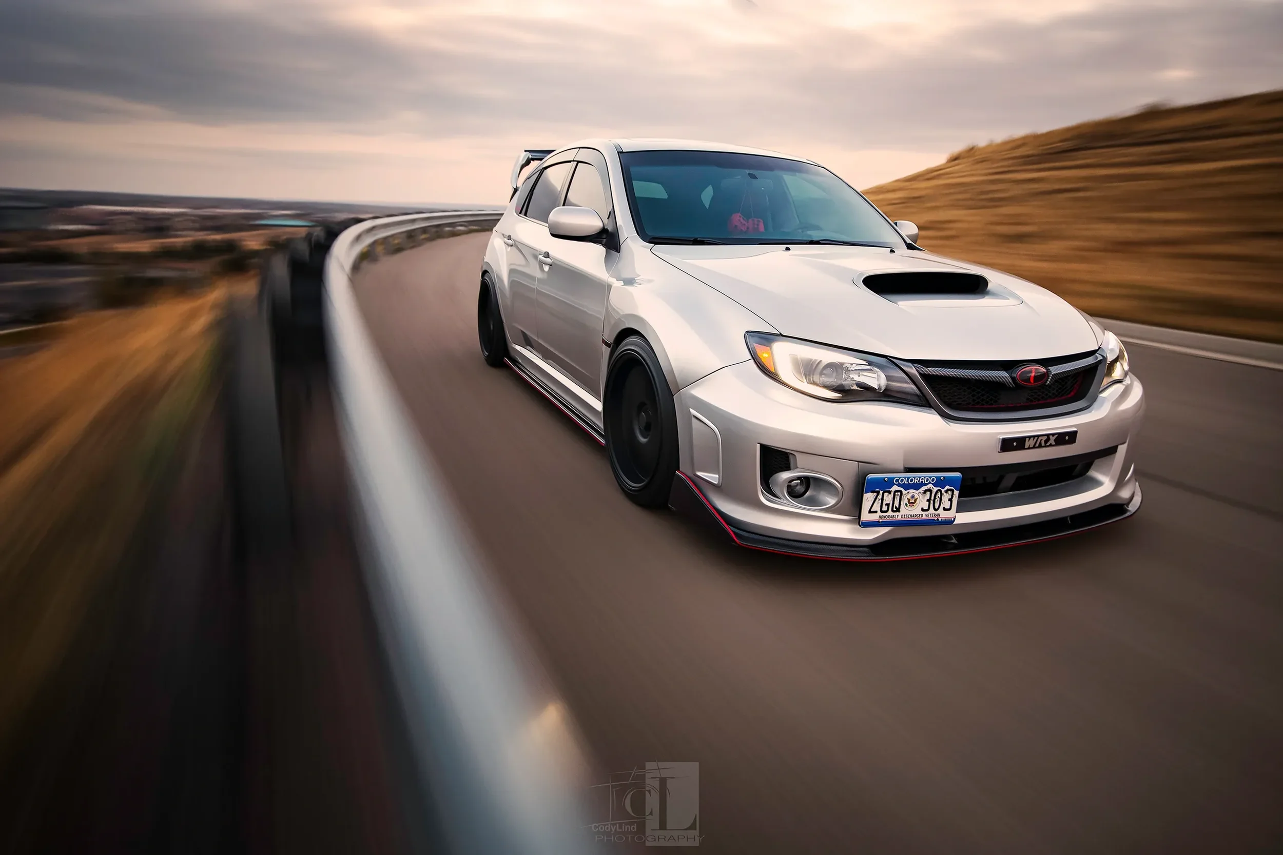 A silver Subaru WRX car with black wheels and a hood scoop is driving on a winding mountain road, captured in motion with a blurred background of hills under a cloudy sky.