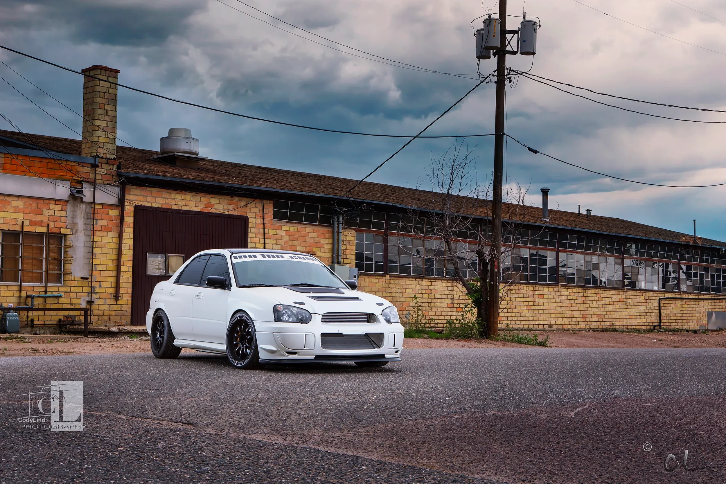 A white modified sedan with black wheels parked on a street in front of a yellow brick building under a cloudy sky.