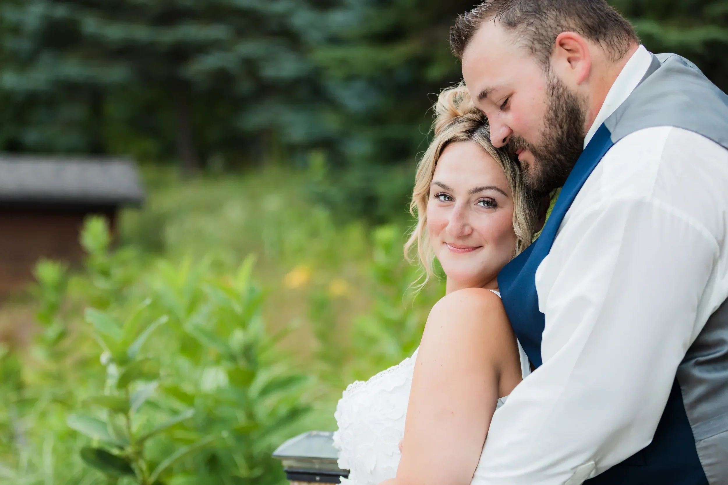 A couple embracing outdoors with greenery in the background, the woman smiling and looking at the camera, the man with his head resting on her head.