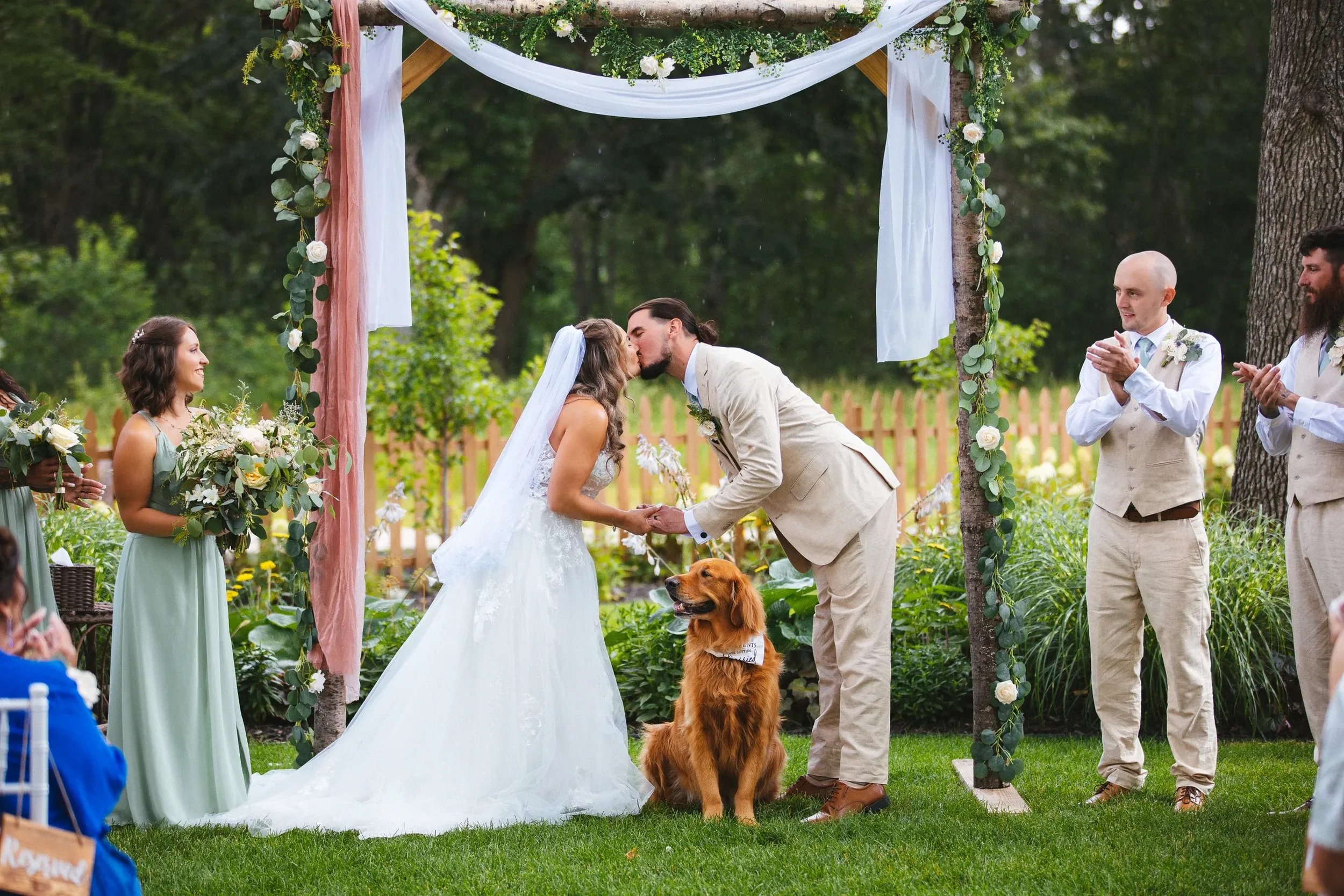 A wedding ceremony outdoors with a bride and groom kissing under an arch decorated with flowers and fabric, surrounded by bridesmaids and groomsmen, with a dog sitting in front of the couple.
