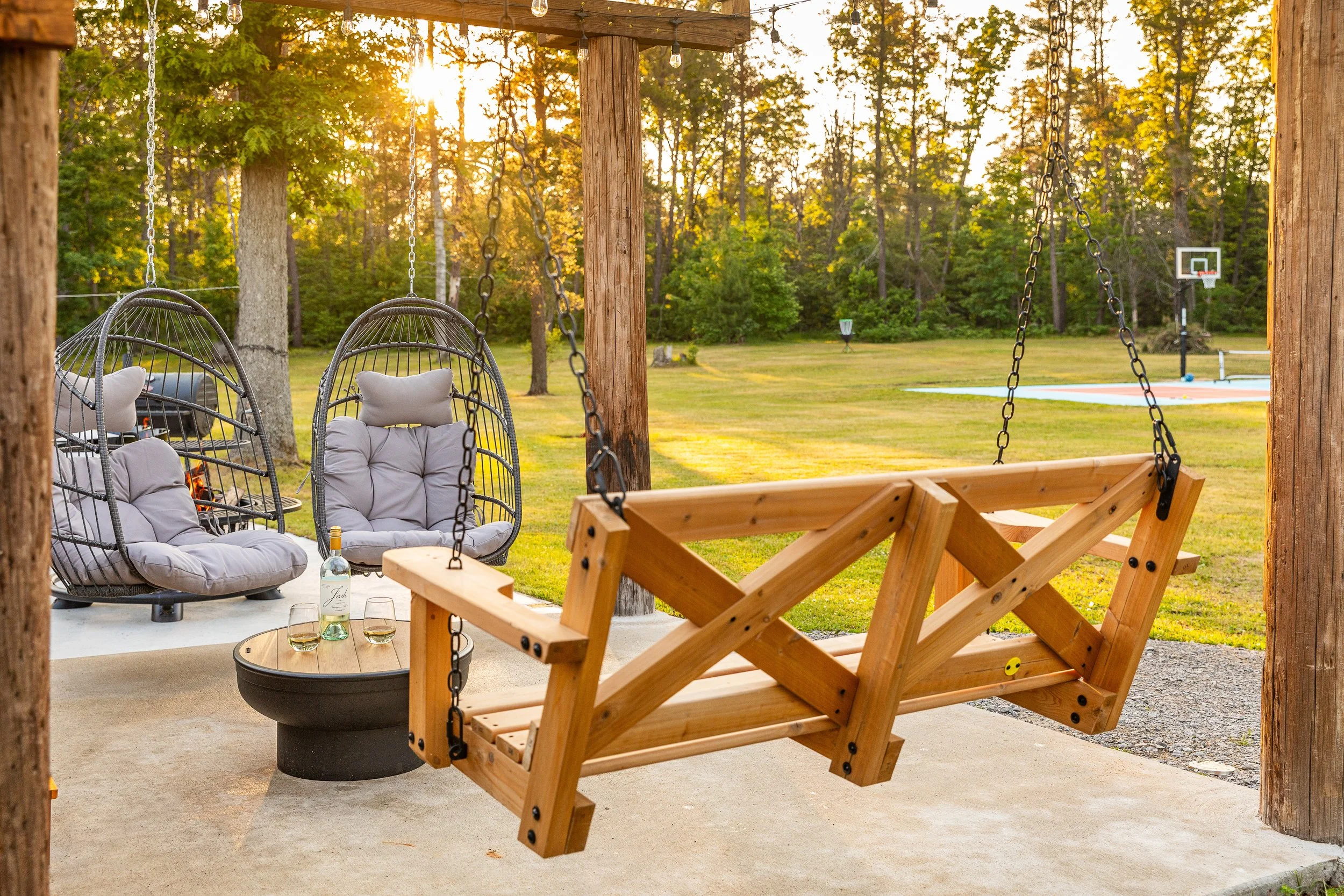 A backyard patio with wooden post and beams, featuring hanging egg chairs with cushions, a wooden porch swing, a round fire pit with wine bottles and glasses, and a grassy yard with a basketball hoop and a small swimming pool in the evening sunlight.