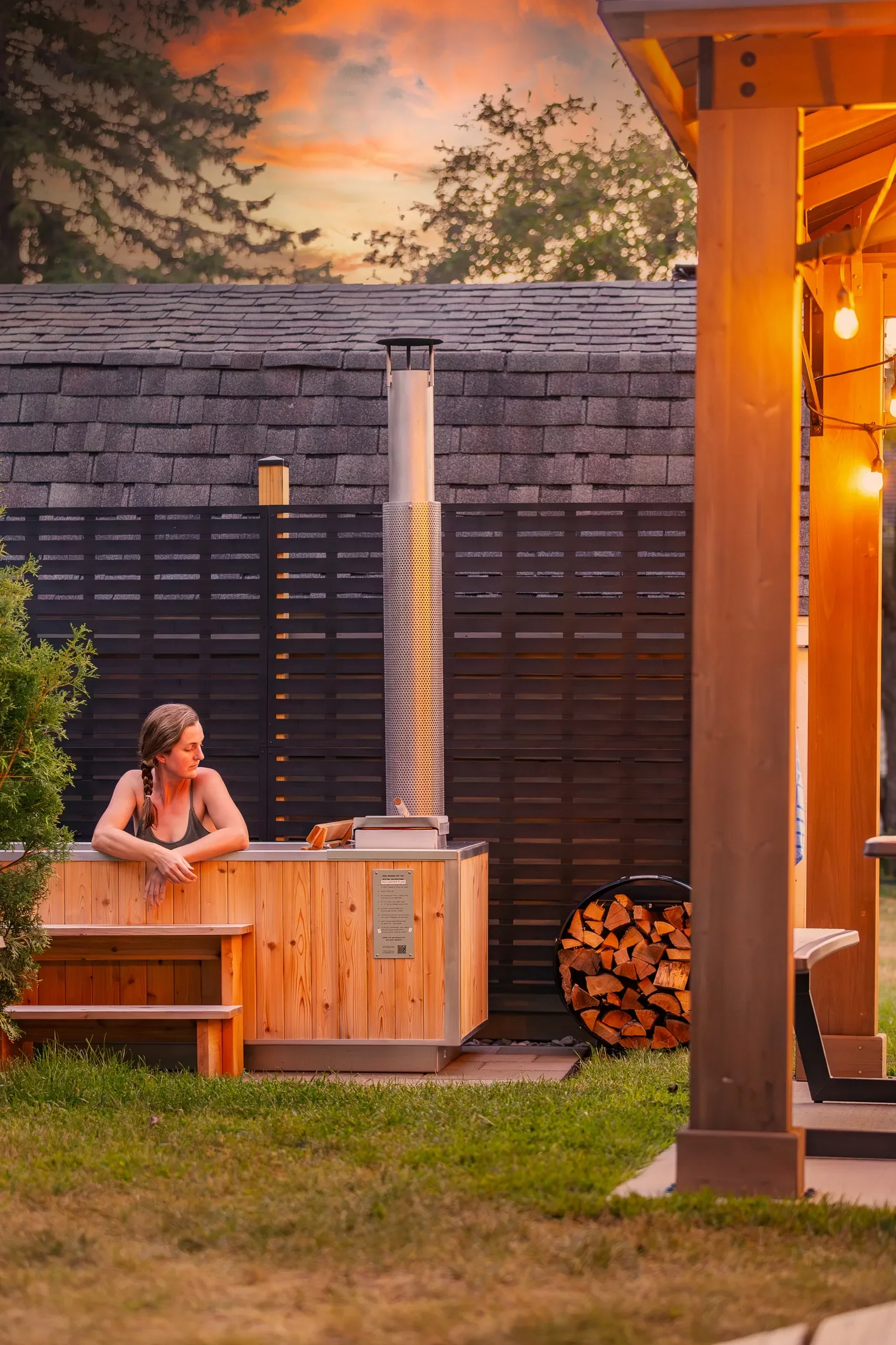 A woman relaxing in an outdoor hot tub at sunset, with a stack of firewood nearby and warm lights illuminating a wooden patio.