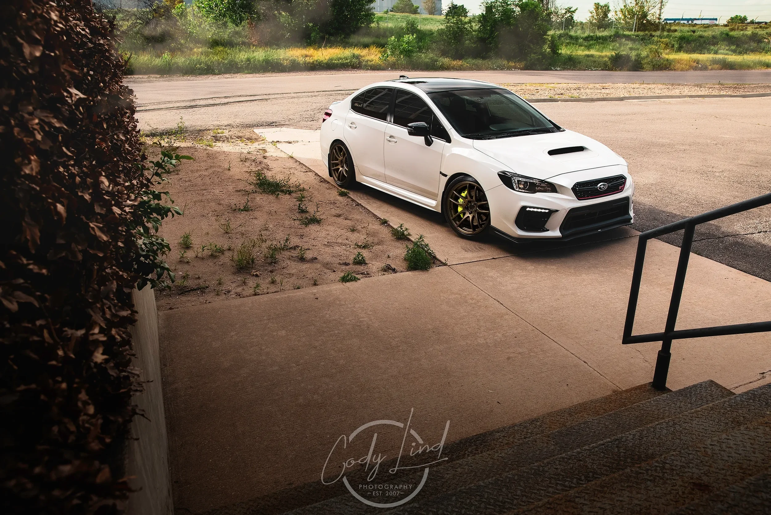 A white Subaru WRX parked on a concrete driveway, with black tinted windows, aftermarket bronze wheels, and lime green brake calipers, near a set of outdoor stairs.