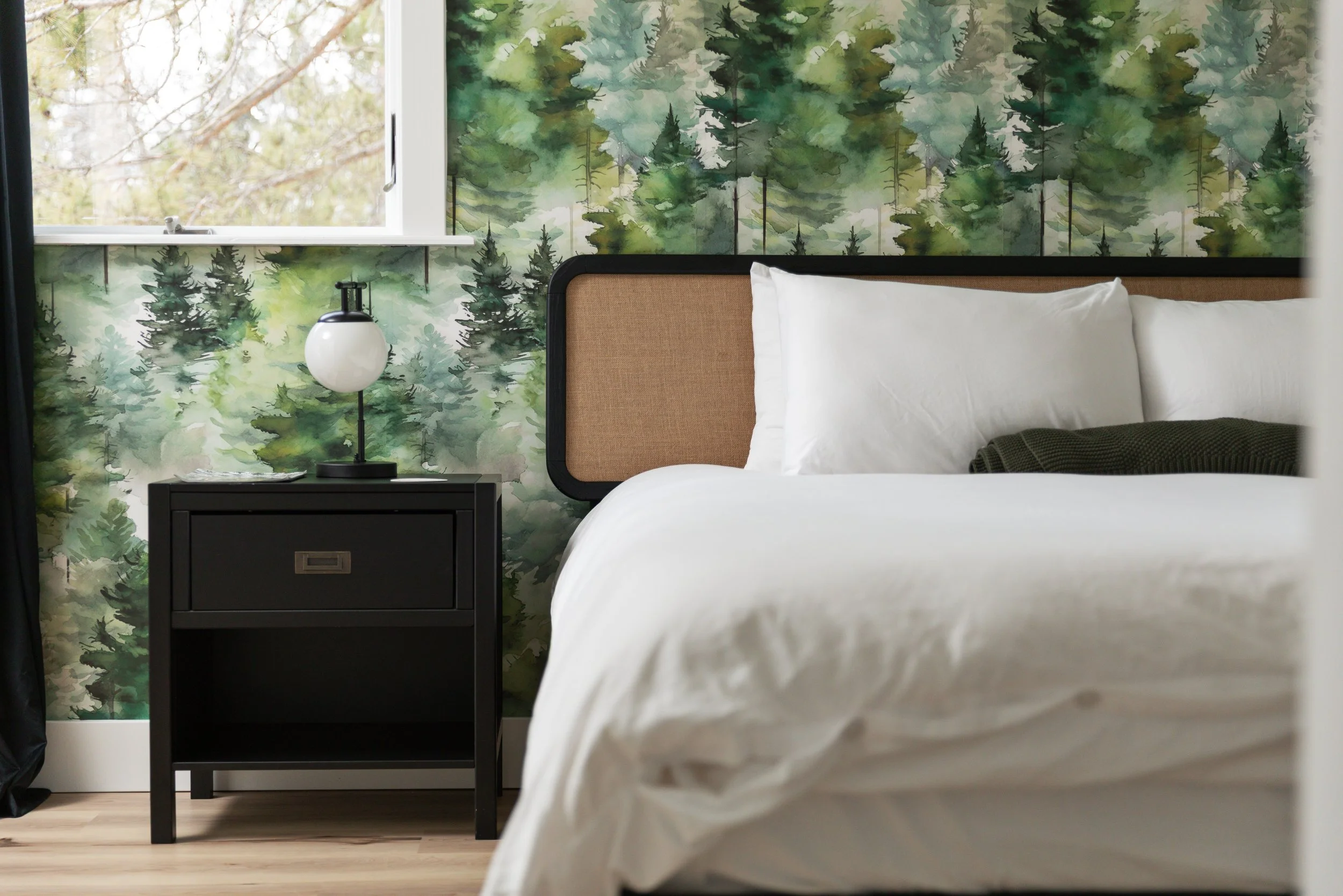 A bedroom with a bed, white bedding, and pillows, a black bedside table with a lamp on it, and wallpaper with a green tree forest pattern behind the bed. A window is partially open to the left, with trees visible outside.