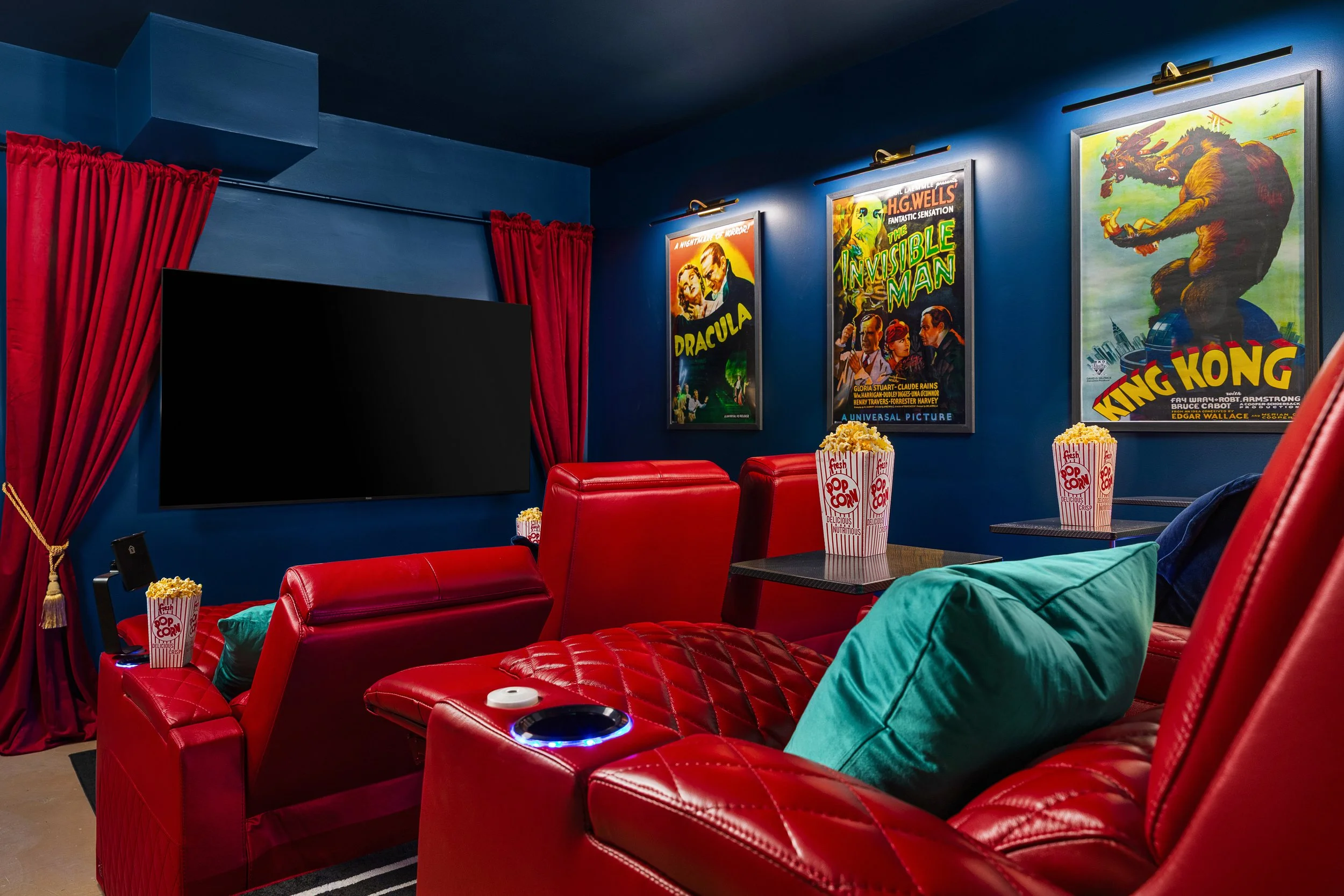 Home theater room with red leather recliners, a large flat-screen TV, vintage horror and monster movie posters on blue walls, and buckets of popcorn on small tables.