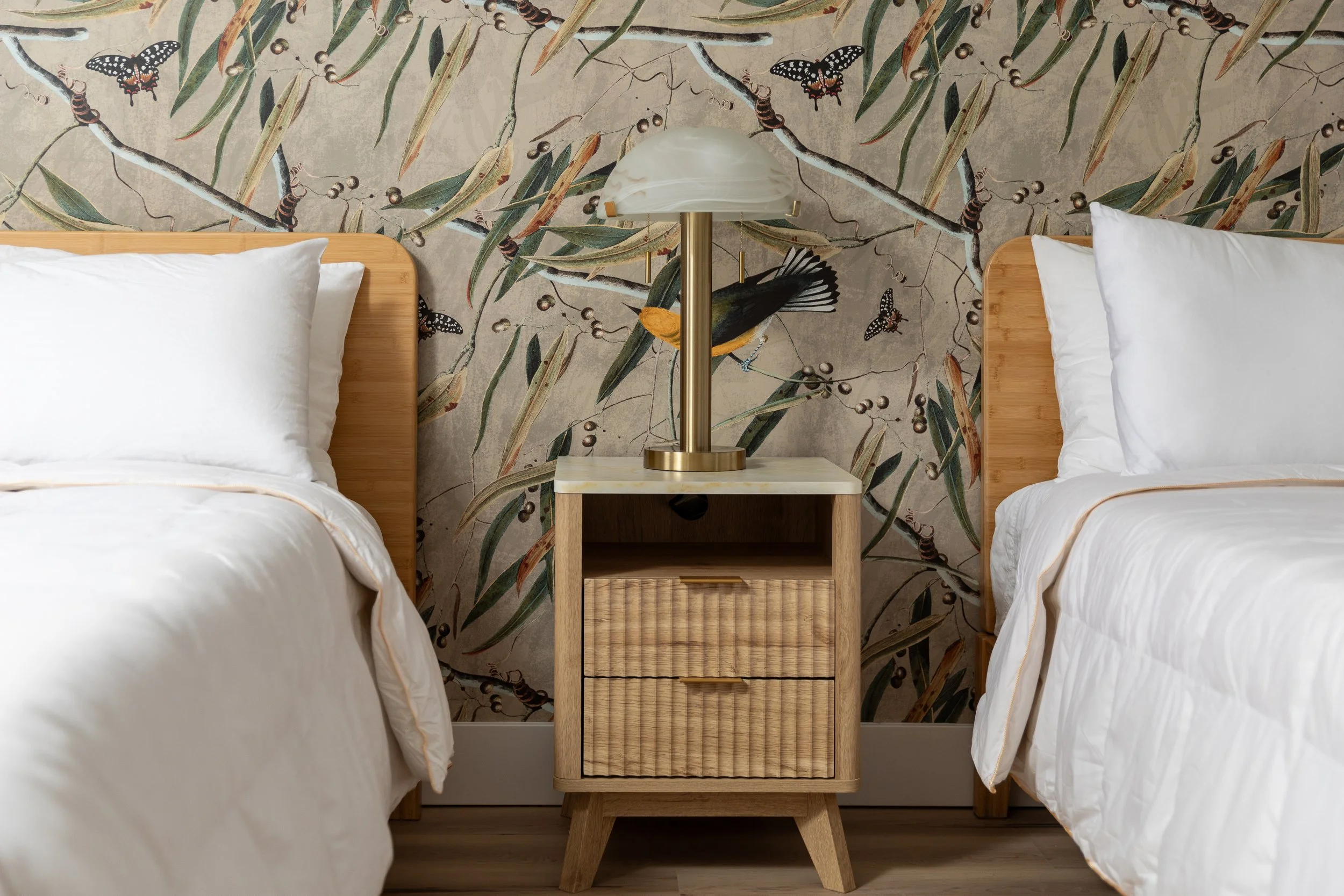A bedroom with two beds, a nightstand in between with a lampshade, and is decorated with floral and bird wallpaper.
