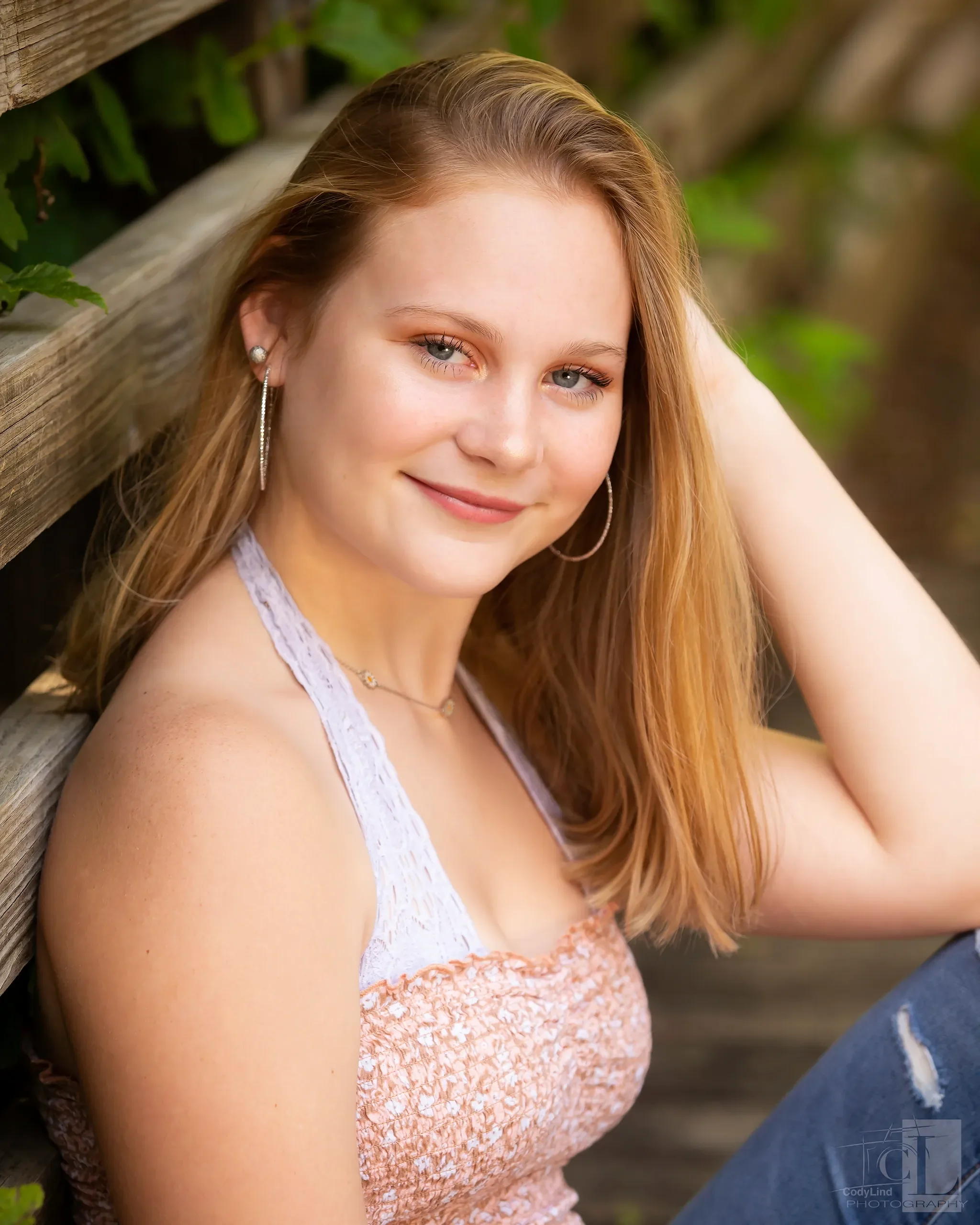 Young woman with red hair and blue eyes smiling, sitting outdoors near a wooden fence with green foliage, wearing a light purple and peach sleeveless top, hoop earrings, and a necklace.